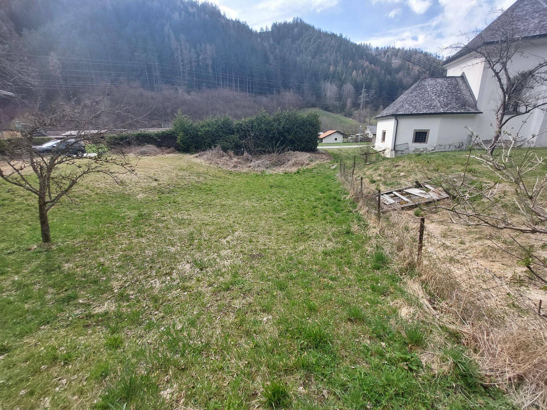 Grünes Grundstück mit Zaun, Kirche und bewaldeten Hügeln im Hintergrund unter blauem Himmel.