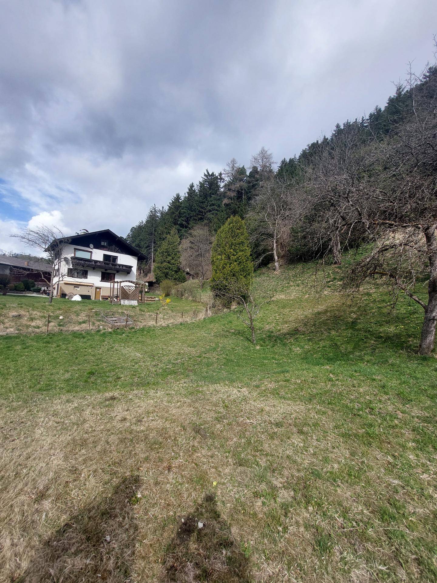 Wohnhaus mit Balkonen an einem grünen Hang, umgeben von bewaldeten Bergen unter bewölktem Himmel.