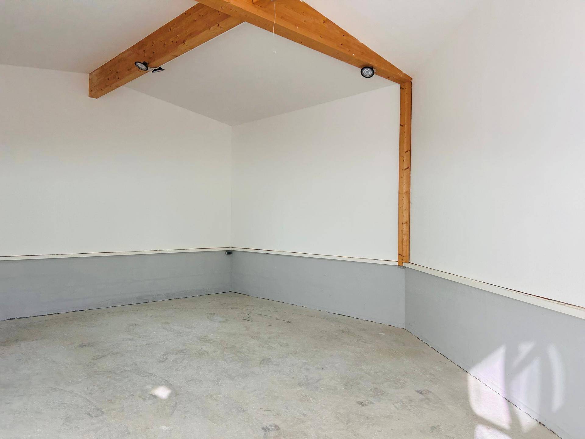 Ecke eines hellen Raumes mit hohen Decken, sichtbaren Holzbalken und einem glatten Betonboden.