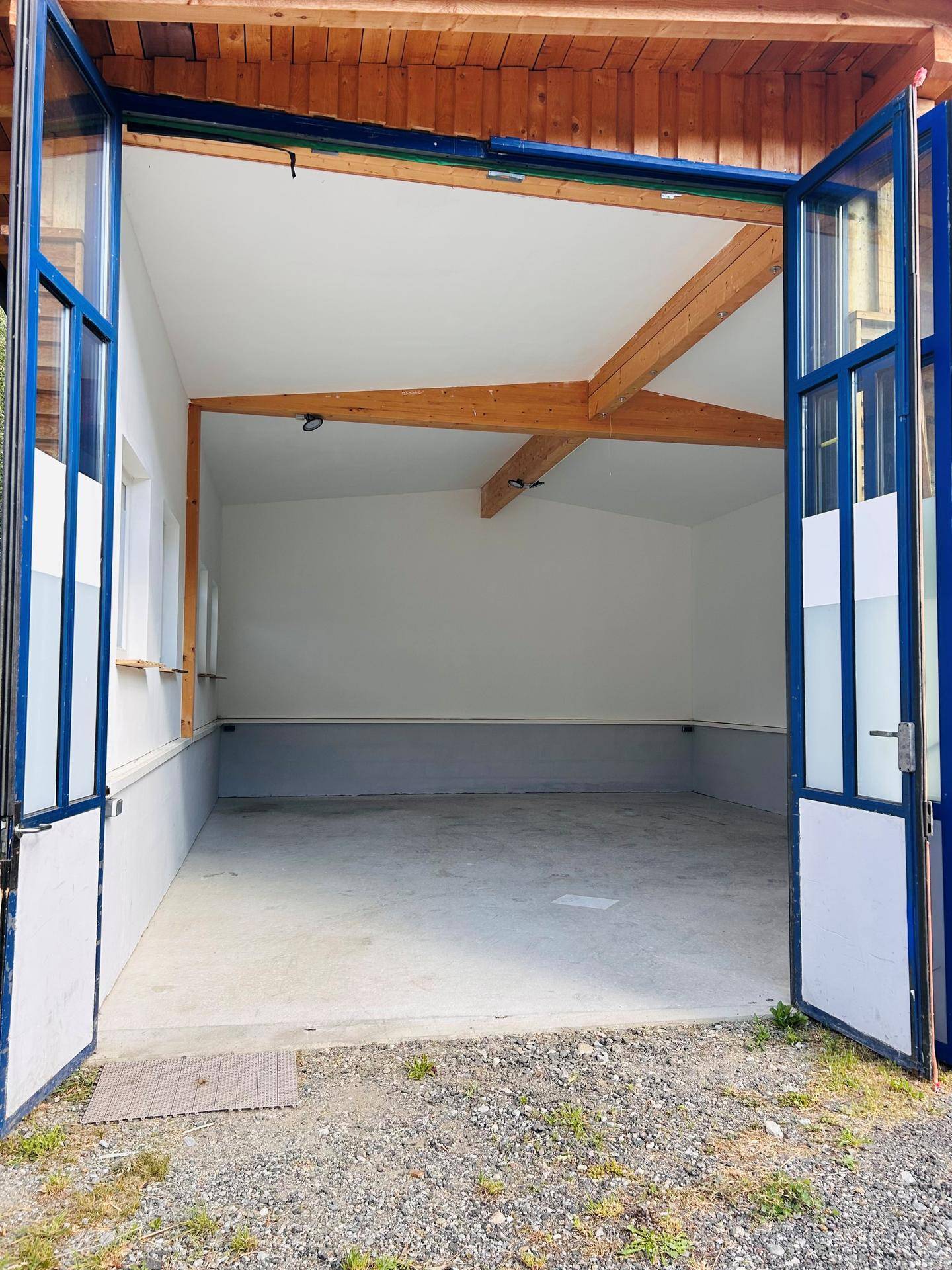 Geräumiger Innenraum mit hohen Decken, sichtbaren Holzbalken und einem Betonboden, zugänglich durch große Falttüren.