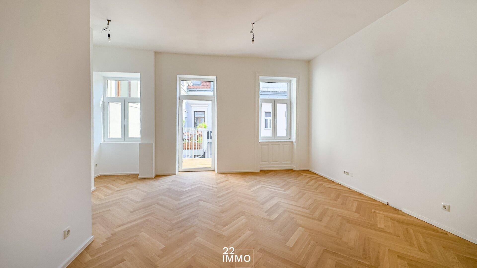 Geräumiges, leeres Wohnzimmer mit Fischgrätparkett und Zugang zum Balkon.