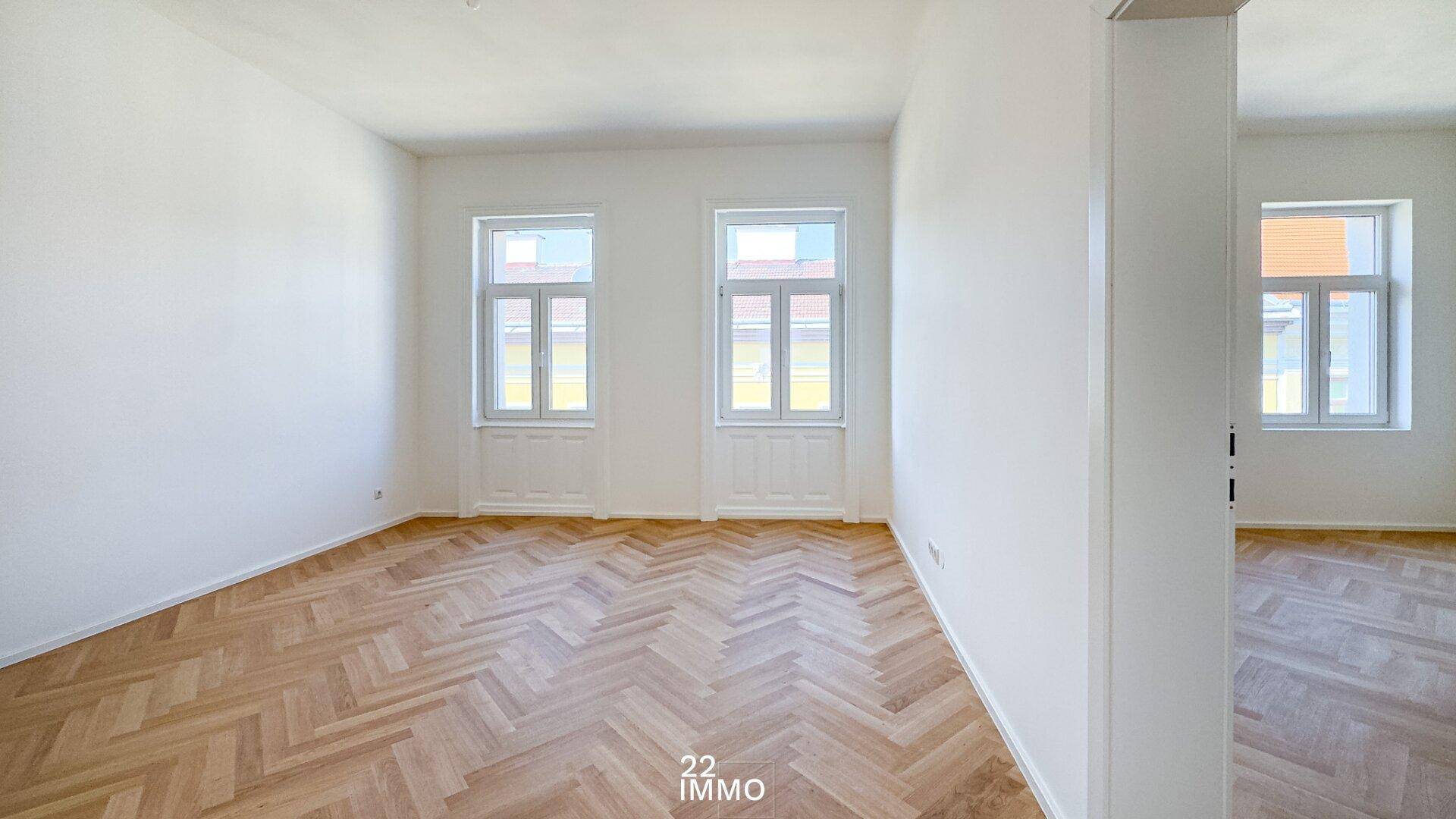 Heller, leerer Raum mit Fischgrätparkett und zwei Fenstern, verbunden mit einem Nebenraum.