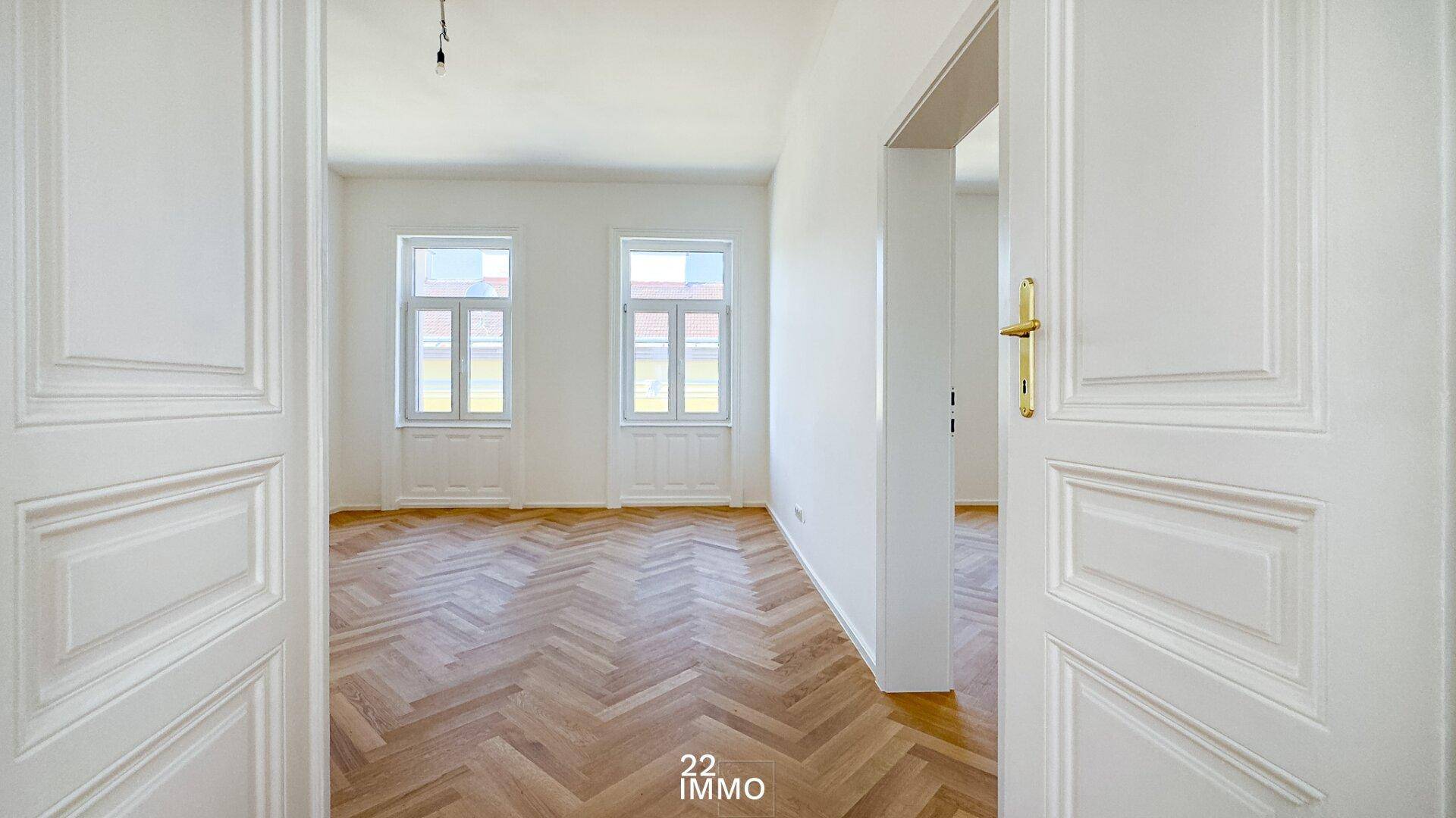 Blick durch offene Türen in einen hellen, leeren Raum mit Fischgrätparkett und Fenstern.