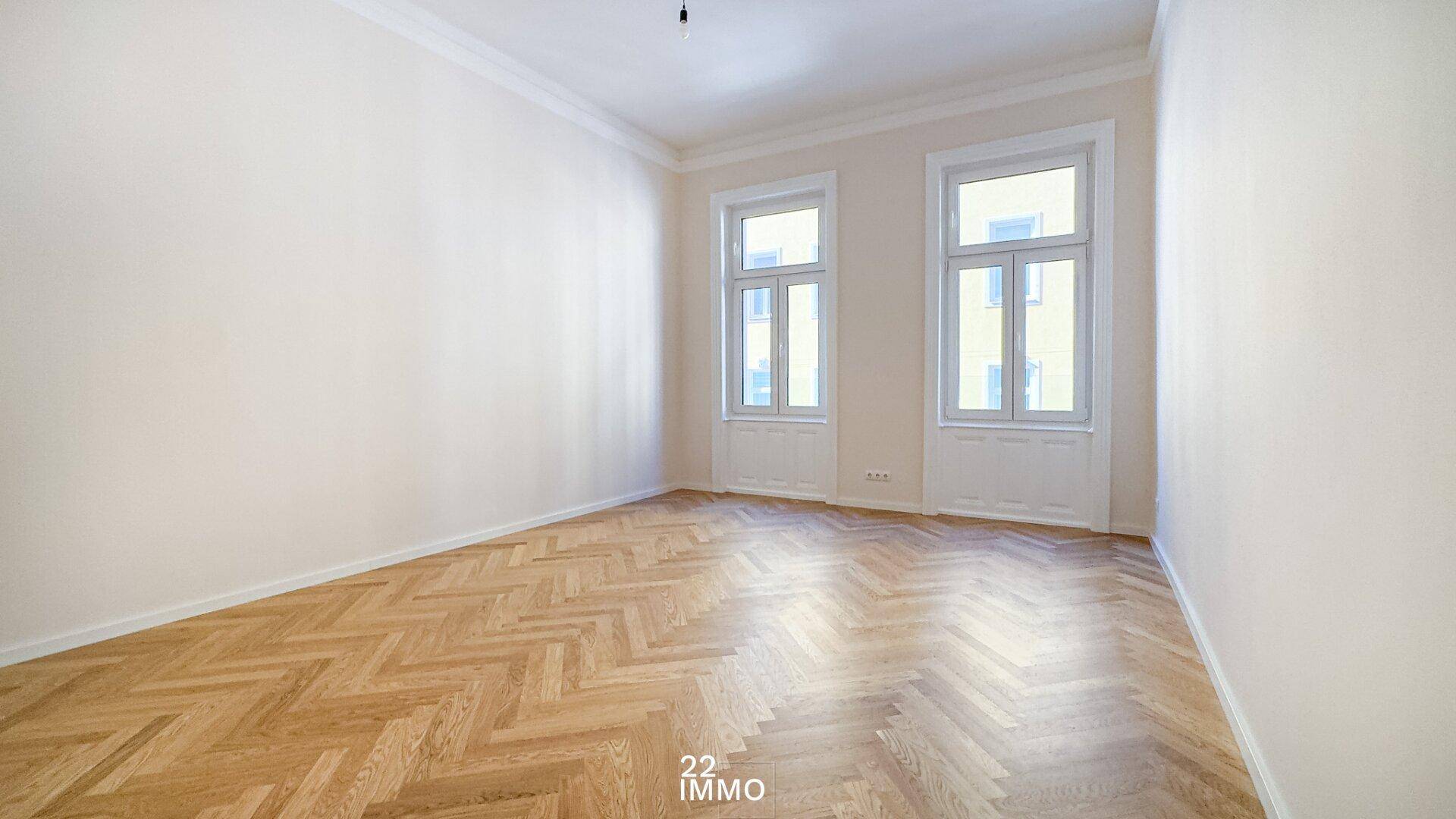 Geräumiges, leeres Zimmer mit Fischgrätparkett und zwei hohen Fenstern, bereit zur individuellen Gestaltung.