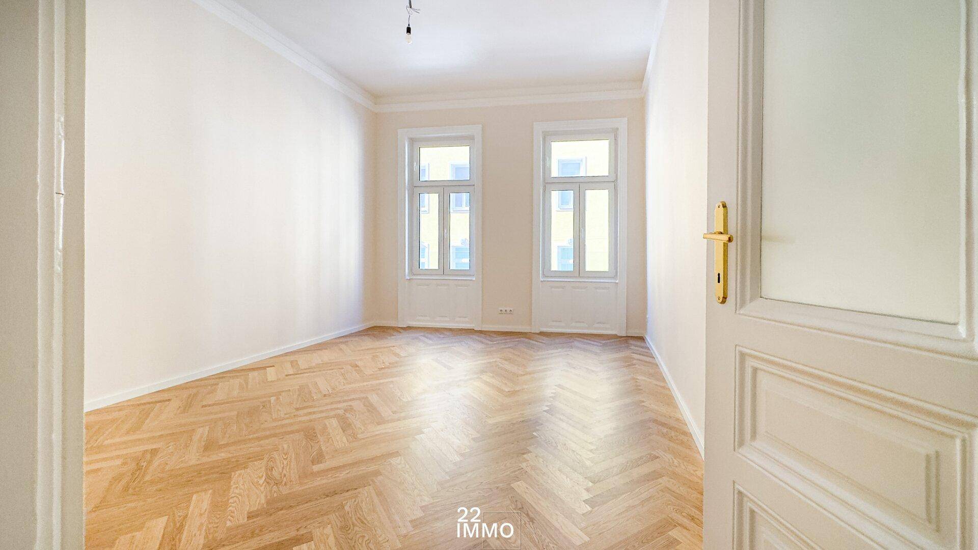 Helles, leeres Zimmer mit Fischgrätparkett und hohen Fenstern, gesehen durch eine offene Tür.