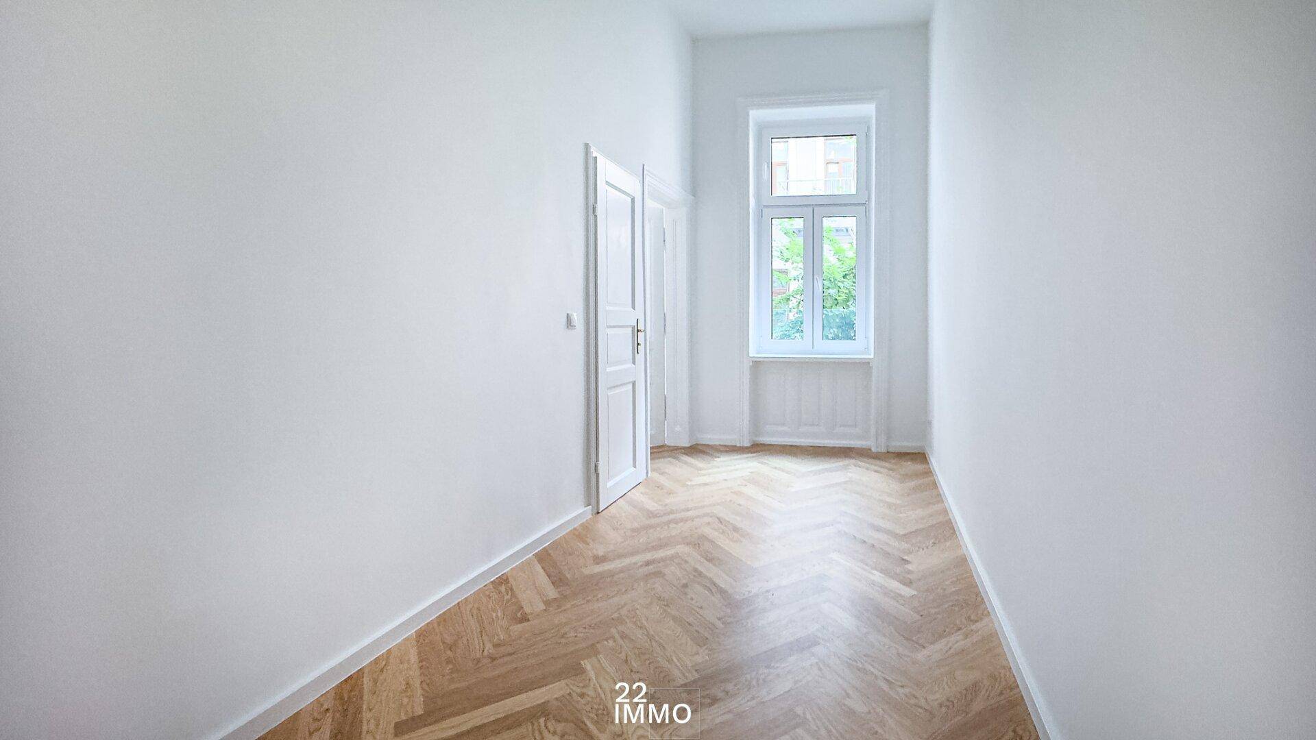 Langes, helles Zimmer mit Fischgrätparkett und einem hohen Fenster, ideal als Schlaf- oder Arbeitszimmer.