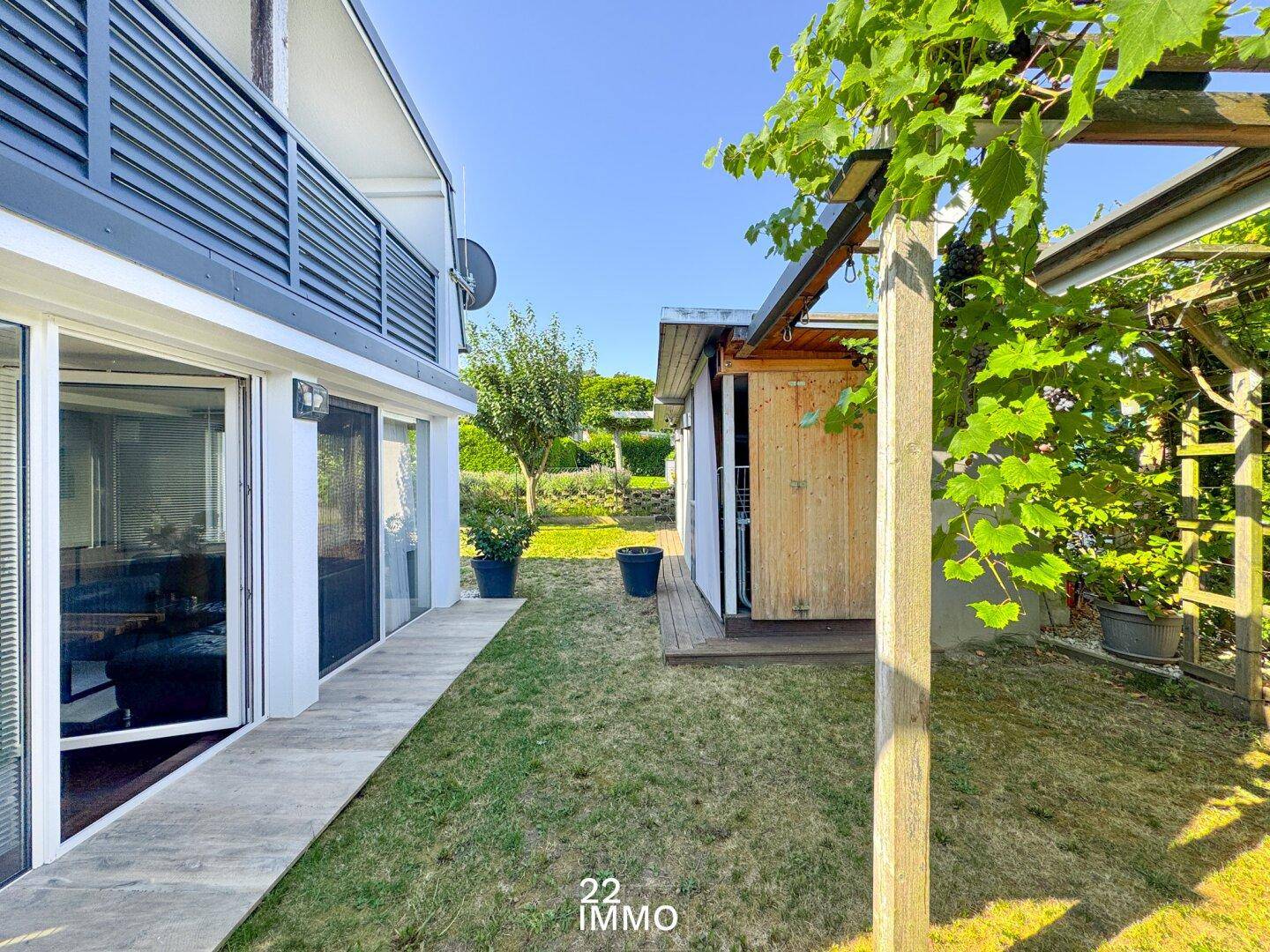 Seitlicher Blick auf das Haus mit großen Fenstern und einem begrünten Gartenbereich mit Pergola.