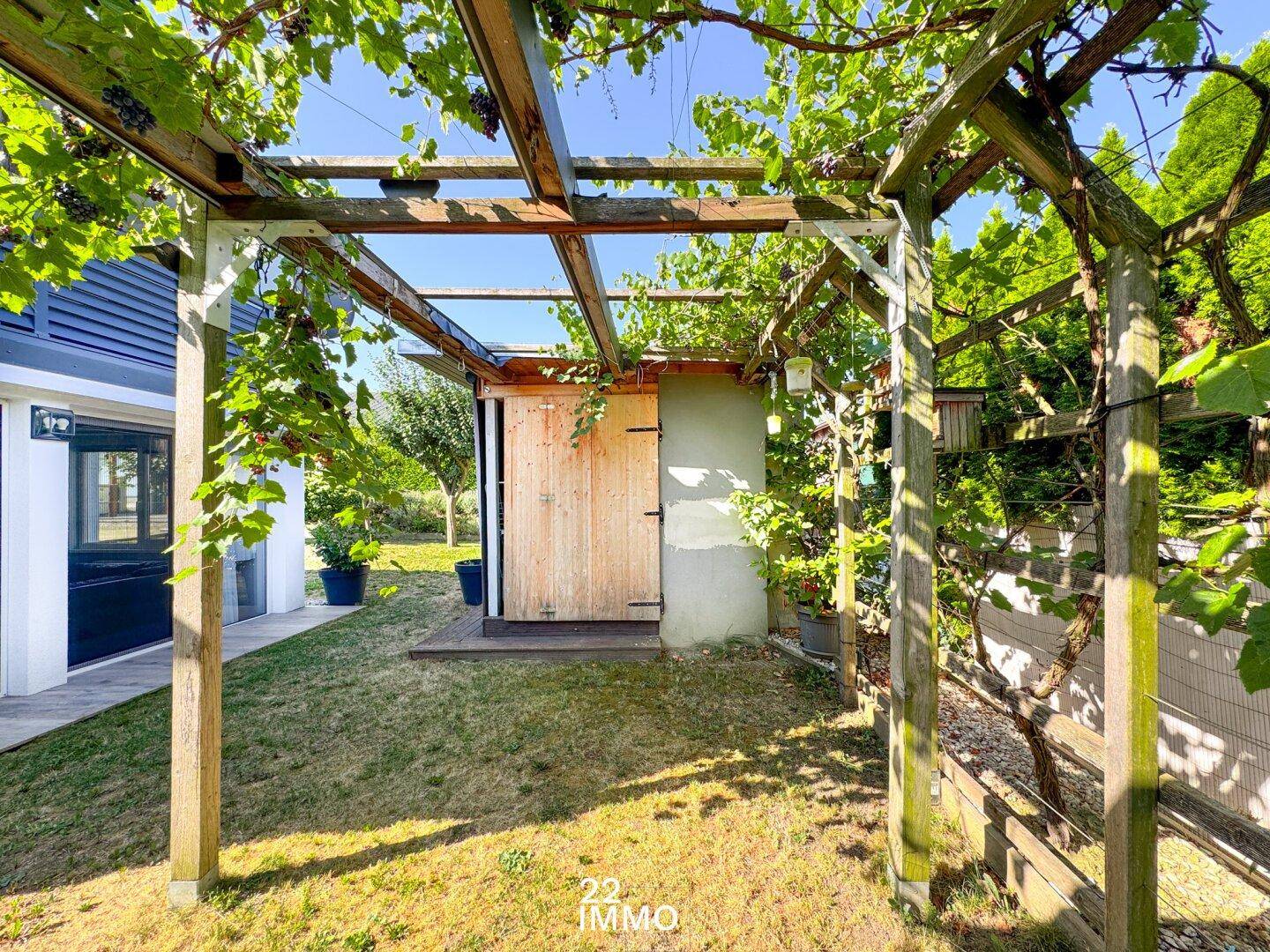 Idyllischer Gartenbereich mit einer Holzpergola und einem kleinen Gartenhaus unter blauem Himmel.