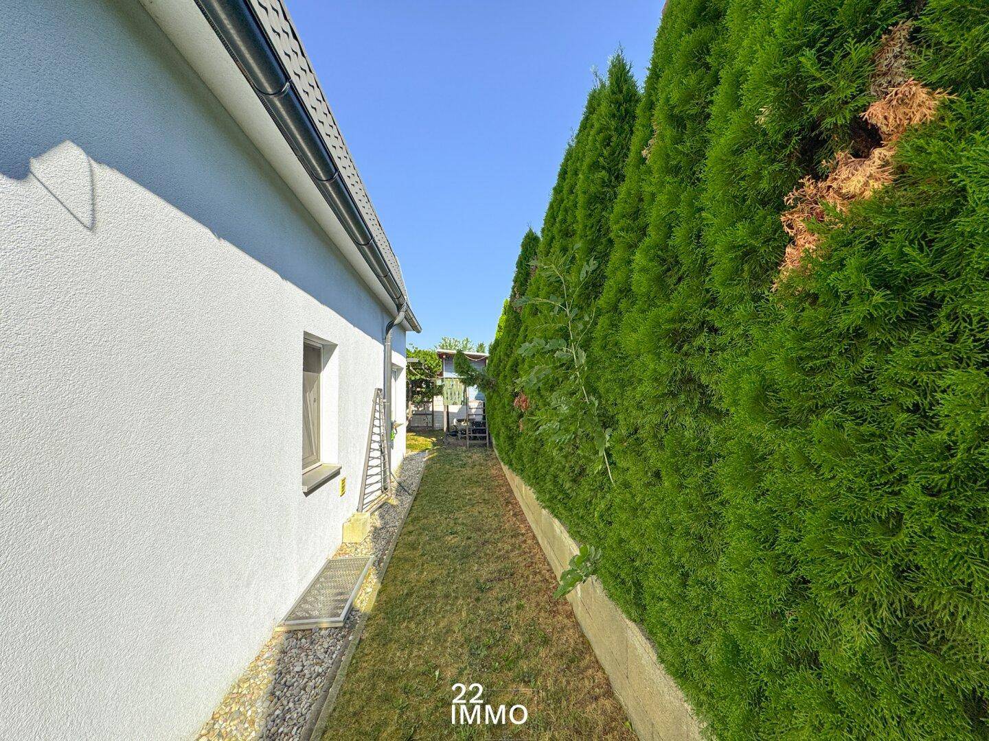 Schmaler Seitenweg entlang des Hauses, begrenzt von einer hohen grünen Hecke und Rasenfläche.