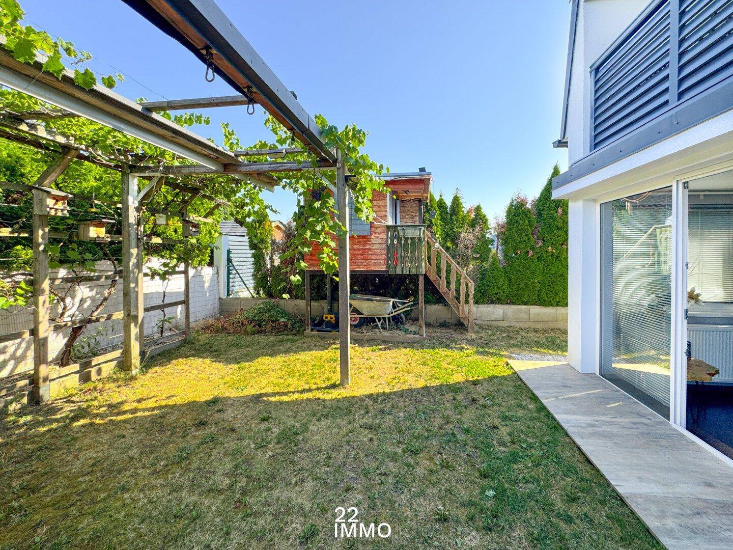 Sonniger Gartenbereich mit Rasenfläche, einer begrünten Pergola und einem Baumhaus, direkt am Haus gelegen.