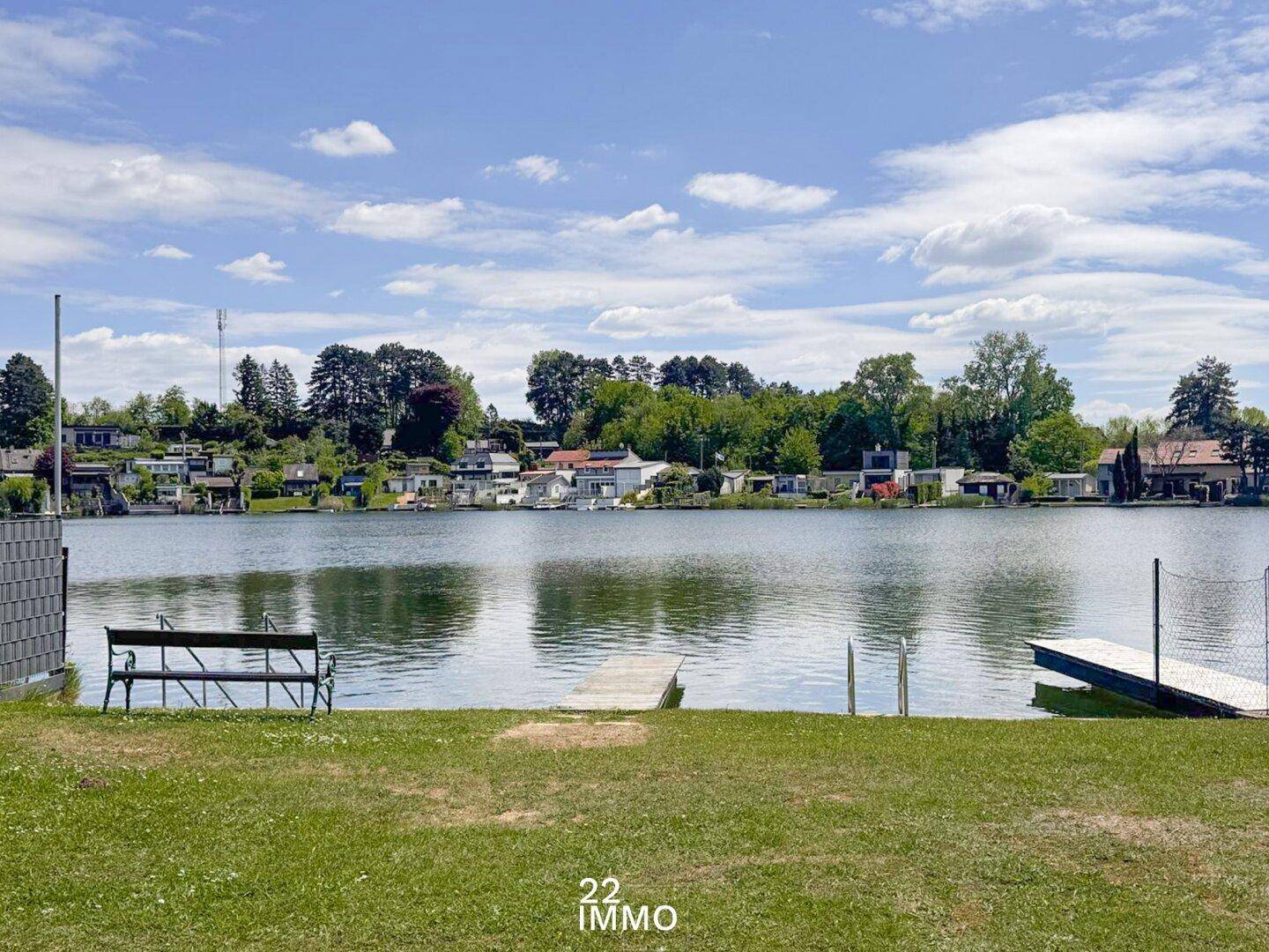 Idyllischer Seeblick mit Steg und Sitzbank unter blauem Himmel und grüner Umgebung.