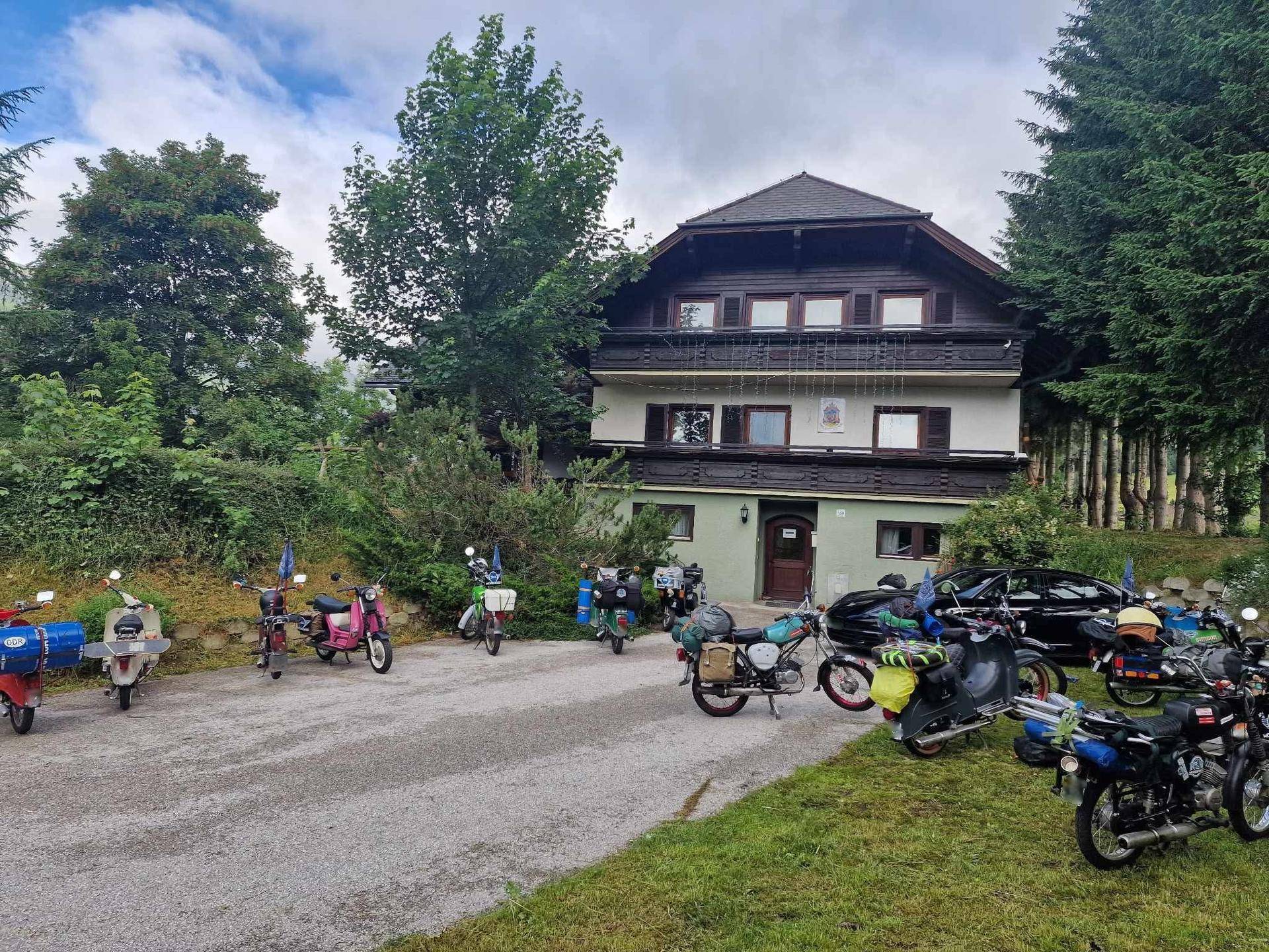 Gebäude am Tag mit zahlreichen Rollern und Motorrädern auf dem Vorplatz, umgeben von grüner Natur.