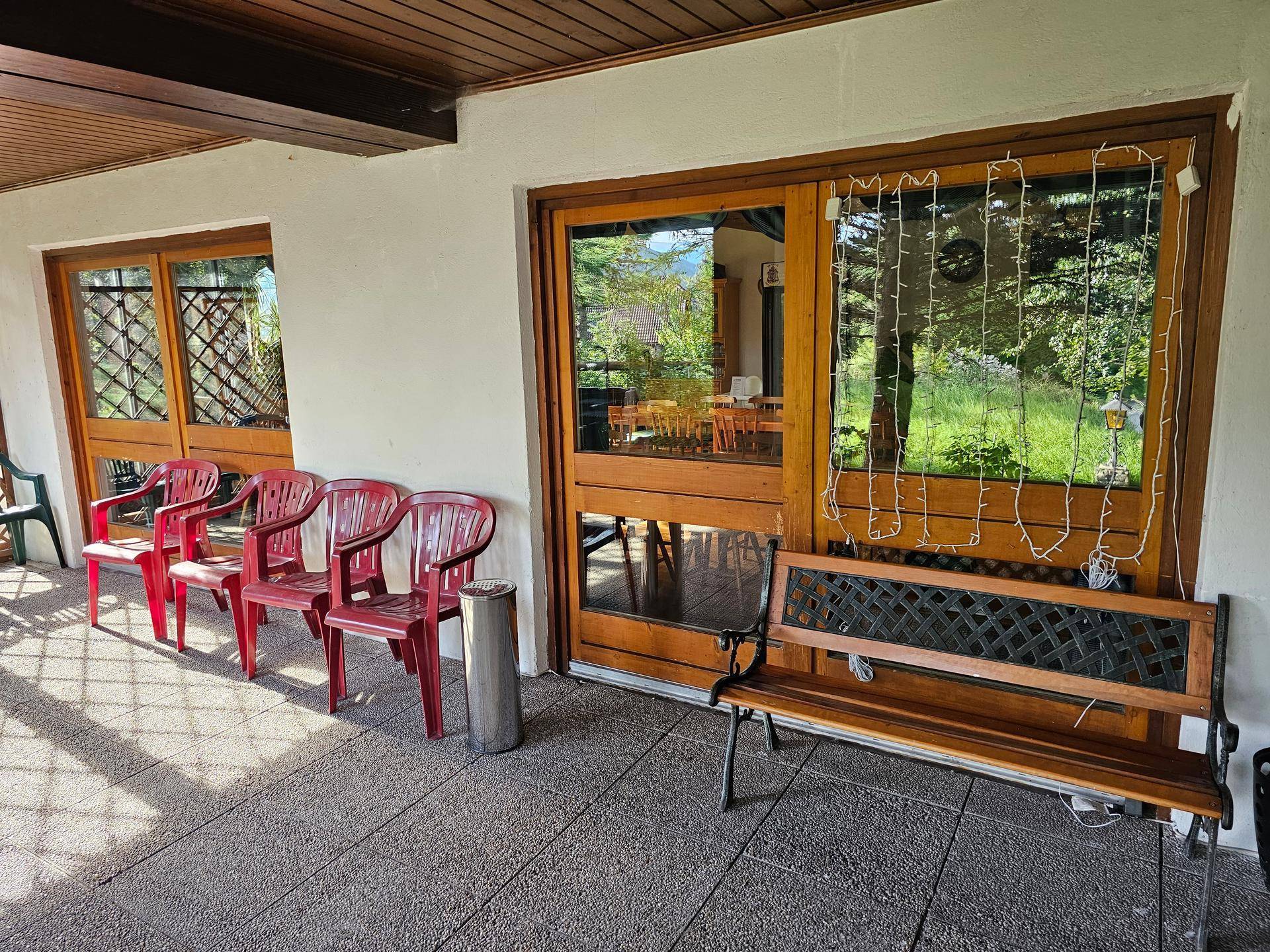 Überdachte Terrasse mit Gartenstühlen und einer Bank, angrenzend an große Glastüren.