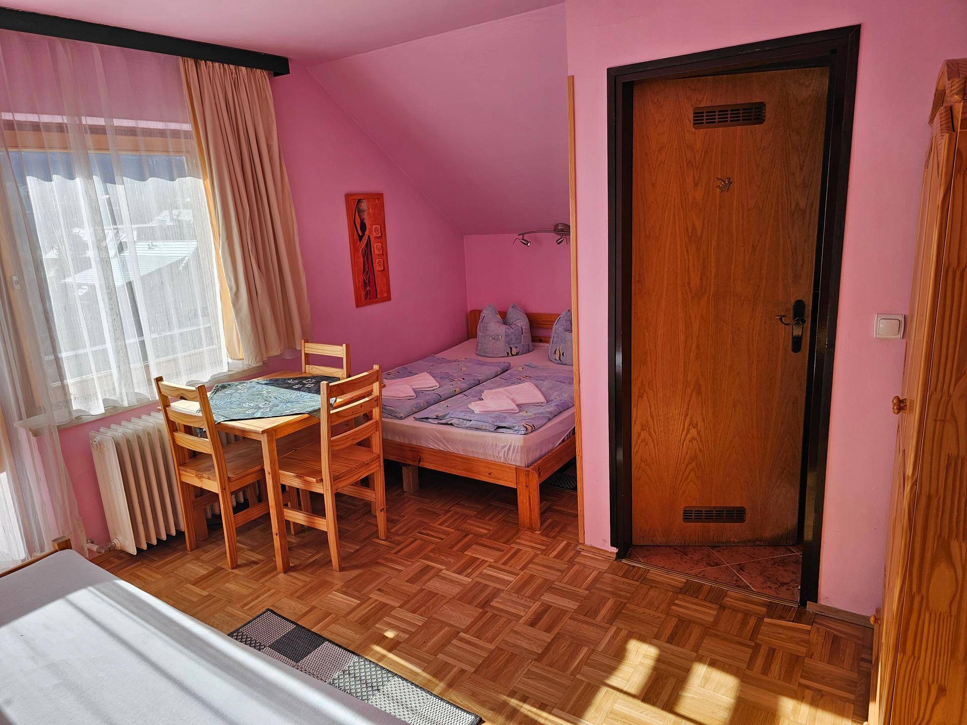 Schlafzimmer mit rosa Wänden, Holzboden und einem kleinen Essbereich neben dem Fenster.
