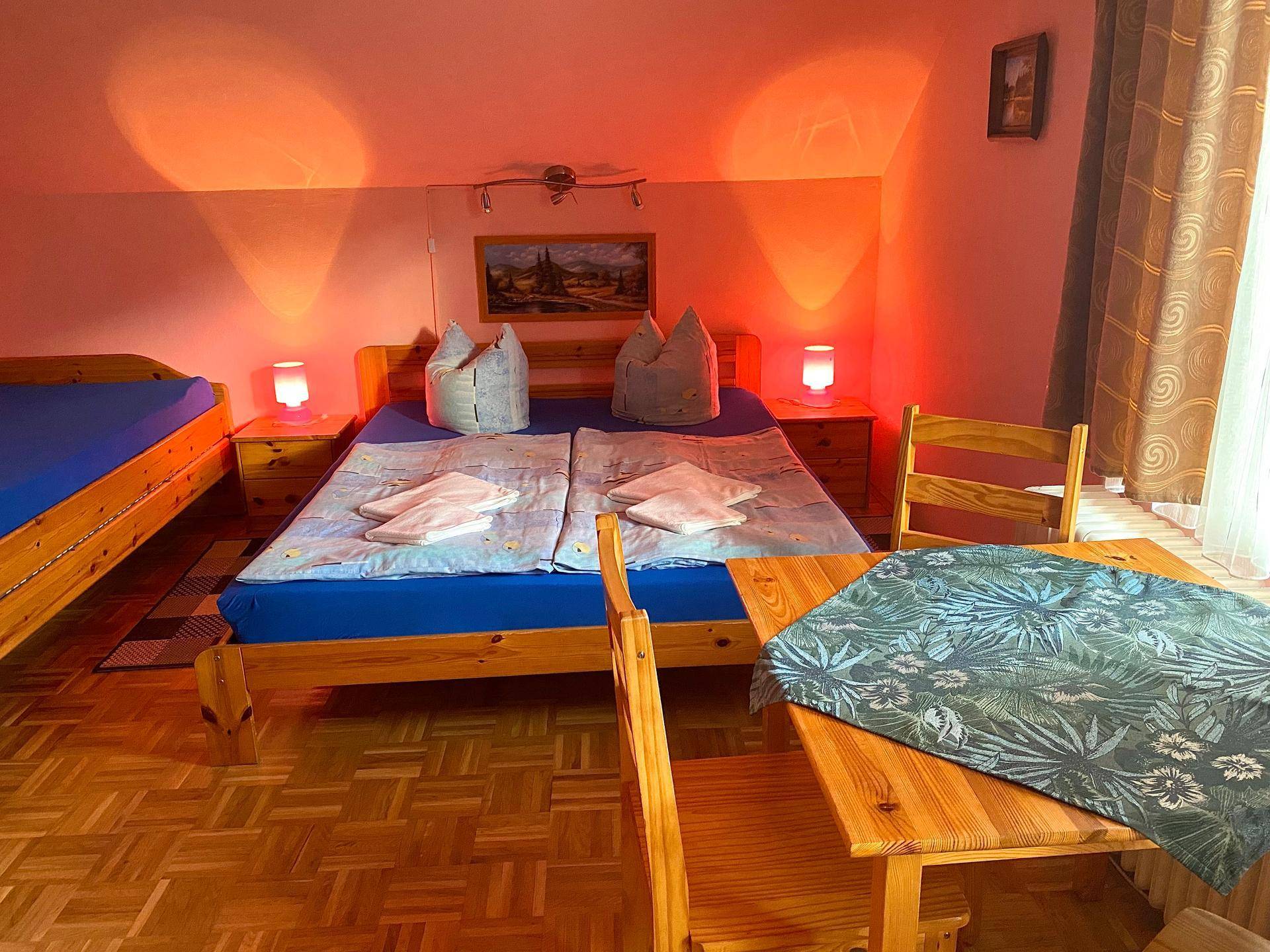 Schlafzimmer mit Doppelbett, Einzelbett und kleinem Tisch, beleuchtet von warmem orangefarbenem Licht.