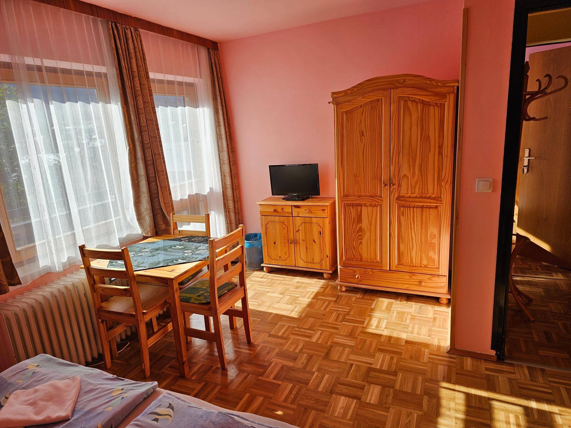 Schlafzimmer mit kleinem Esstisch, Kleiderschrank und Fernseher, in rosa Farbtönen gehalten.