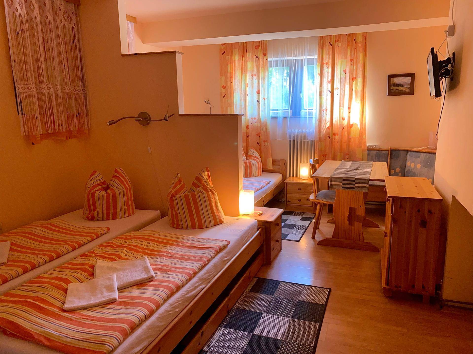 Schlafzimmer mit zwei Einzelbetten, einem kleinen Esstisch und einem Fenster mit orangefarbenen Vorhängen.