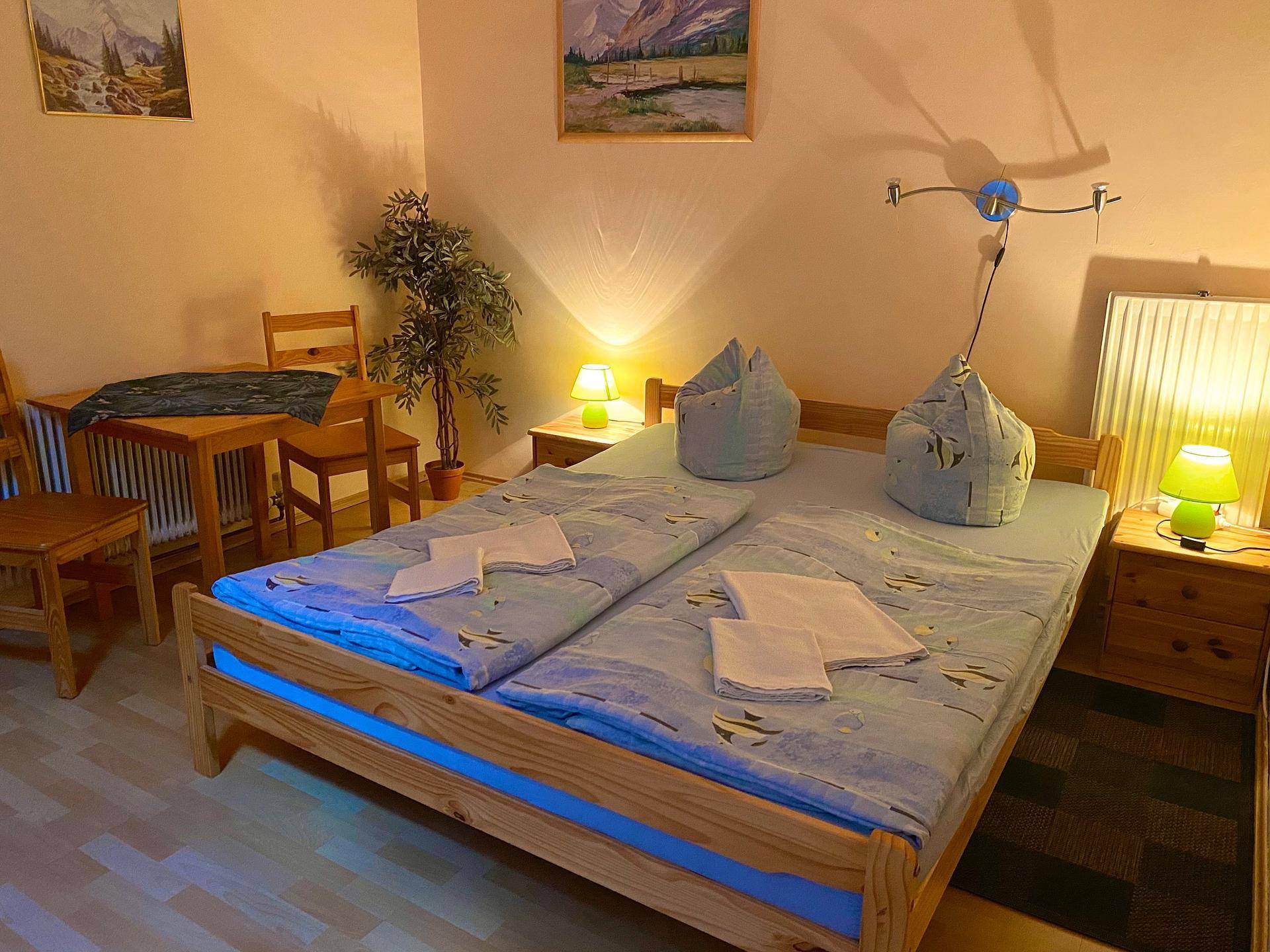 Schlafzimmer mit Doppelbett, kleinem Tisch und einer Pflanze, ausgestattet mit blauer Bettwäsche.
