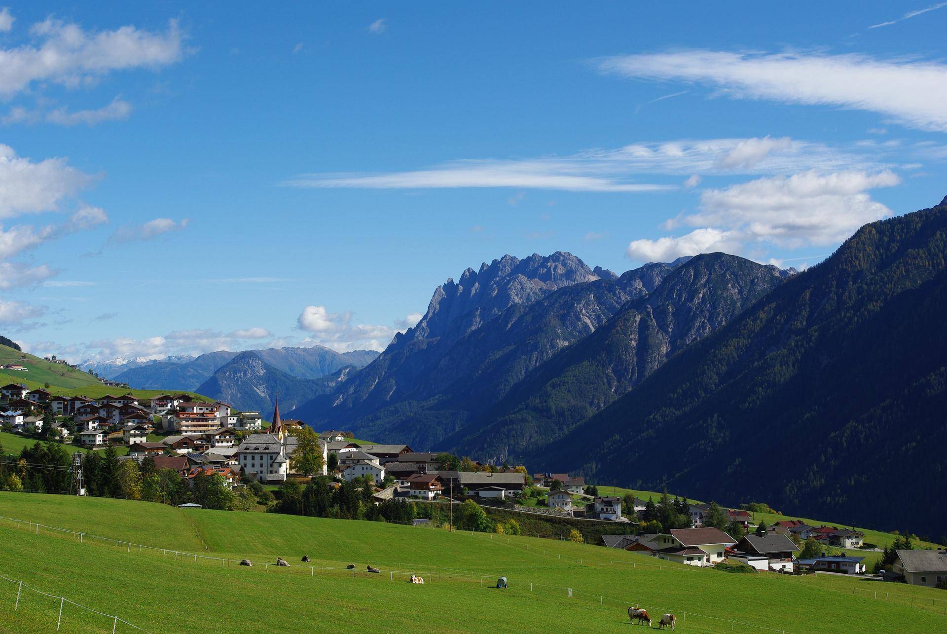 Malerisches Dorf in einem grünen Tal mit majestätischen Bergen unter blauem Himmel.