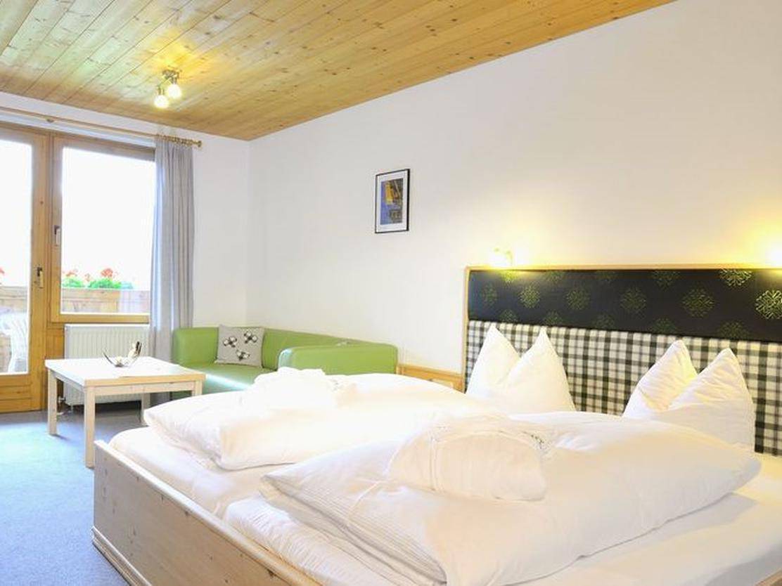 Helles Schlafzimmer mit Doppelbett, Holzdecke und einem grünen Sofa, mit Zugang zu einem Balkon.