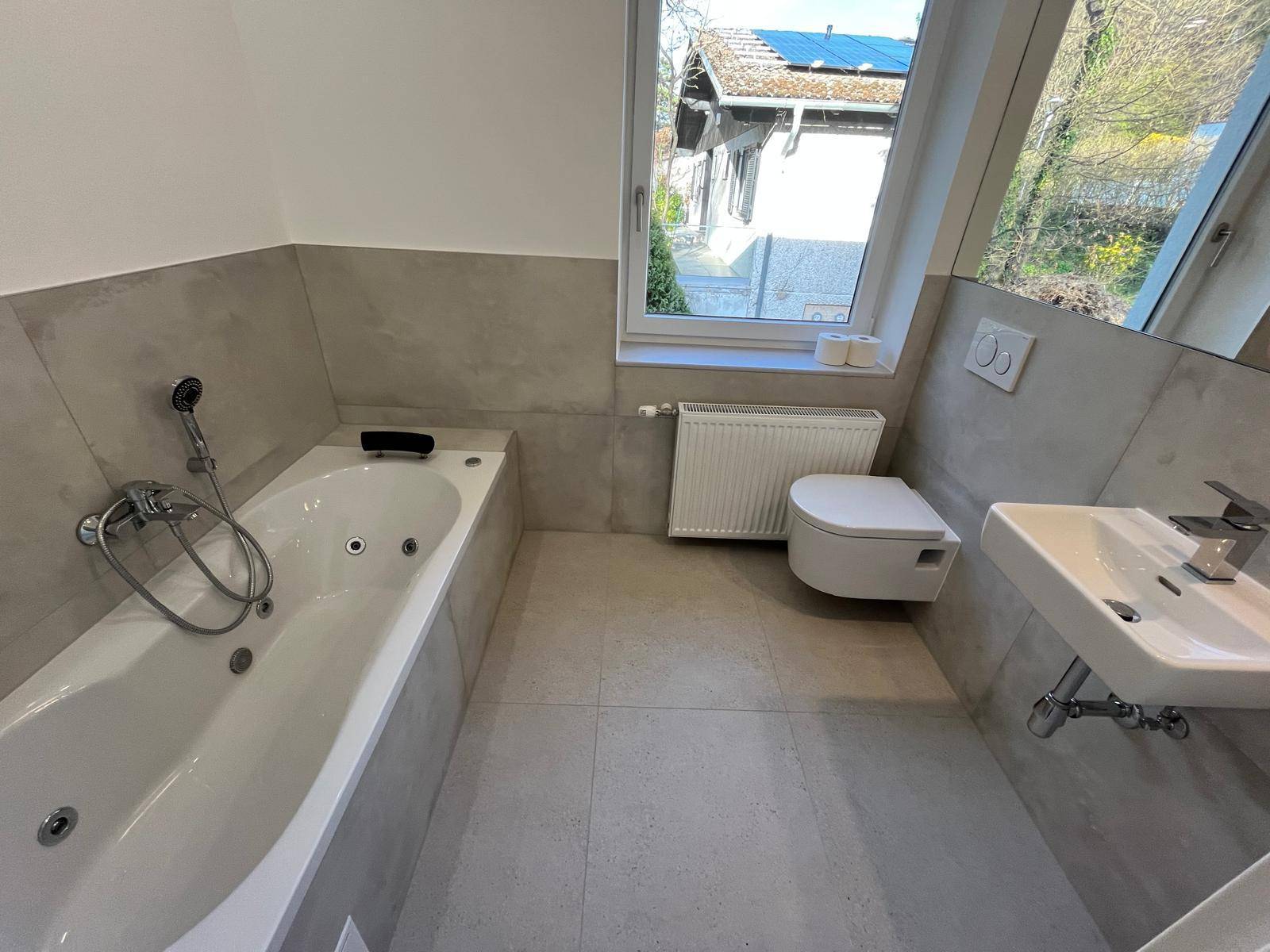 Helles Badezimmer mit Badewanne, Wand-WC und Fenster mit Blick nach draußen.