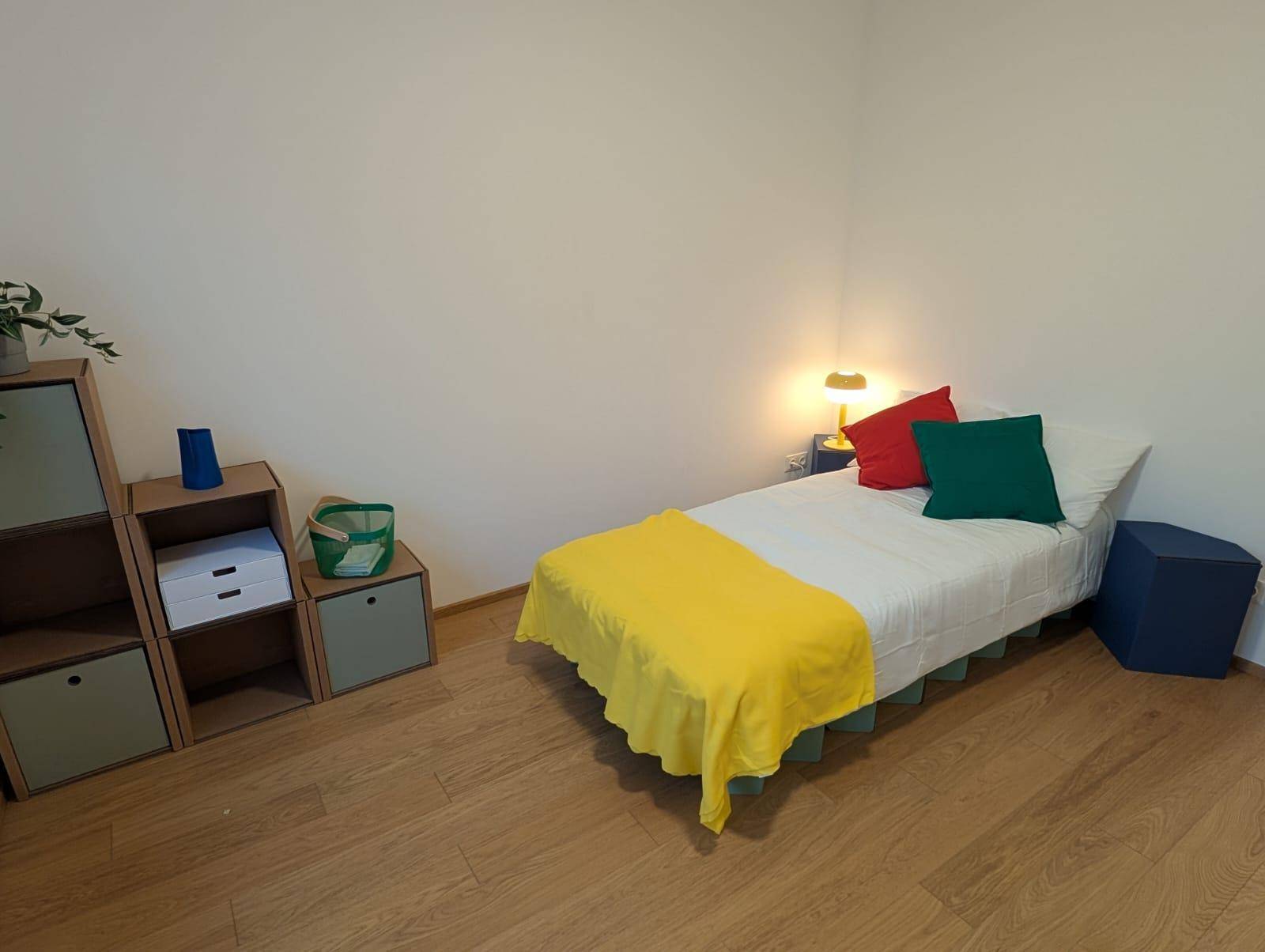 Helles Schlafzimmer mit Holzboden, modernem Regal und farbenfroher Bettwäsche.