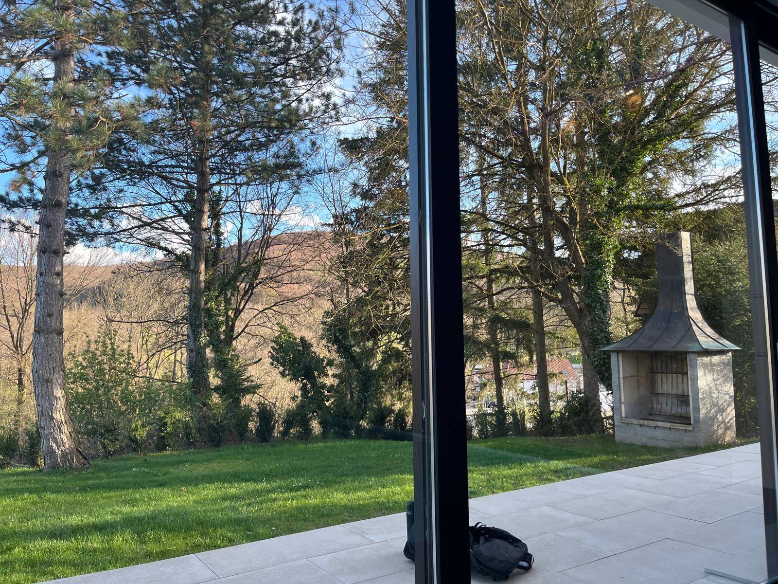 Blick vom Innenraum auf den Garten mit Außengrill und hohen Bäumen.