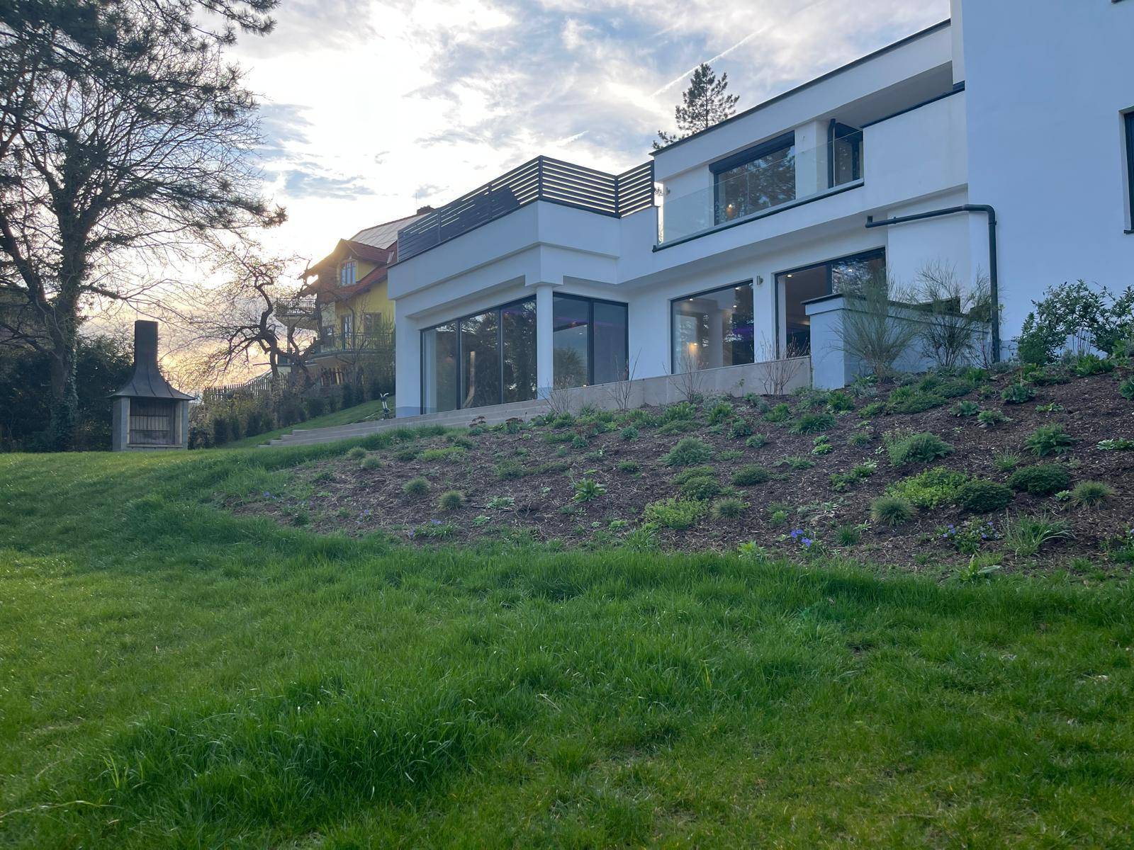 Gartenbereich der modernen Villa mit Rasenfläche, Bepflanzung und einem Außengrill.