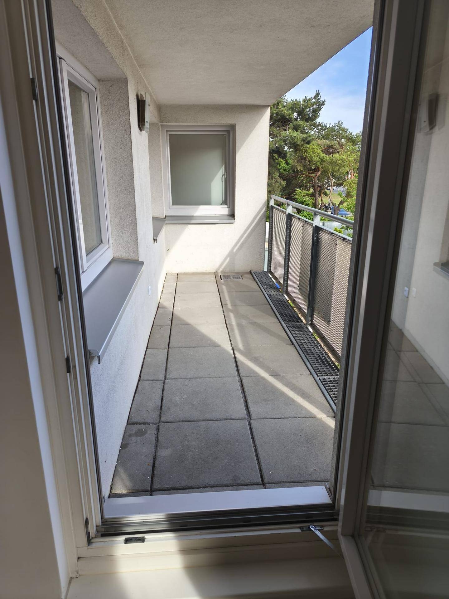 Heller Balkon mit Fliesenboden und Metallgeländer, umgeben von grünen Bäumen.