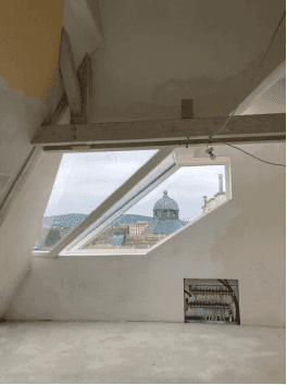 Dachgeschossraum im Bau mit Dachfenster und Blick auf die umliegenden Dächer und Kuppel.