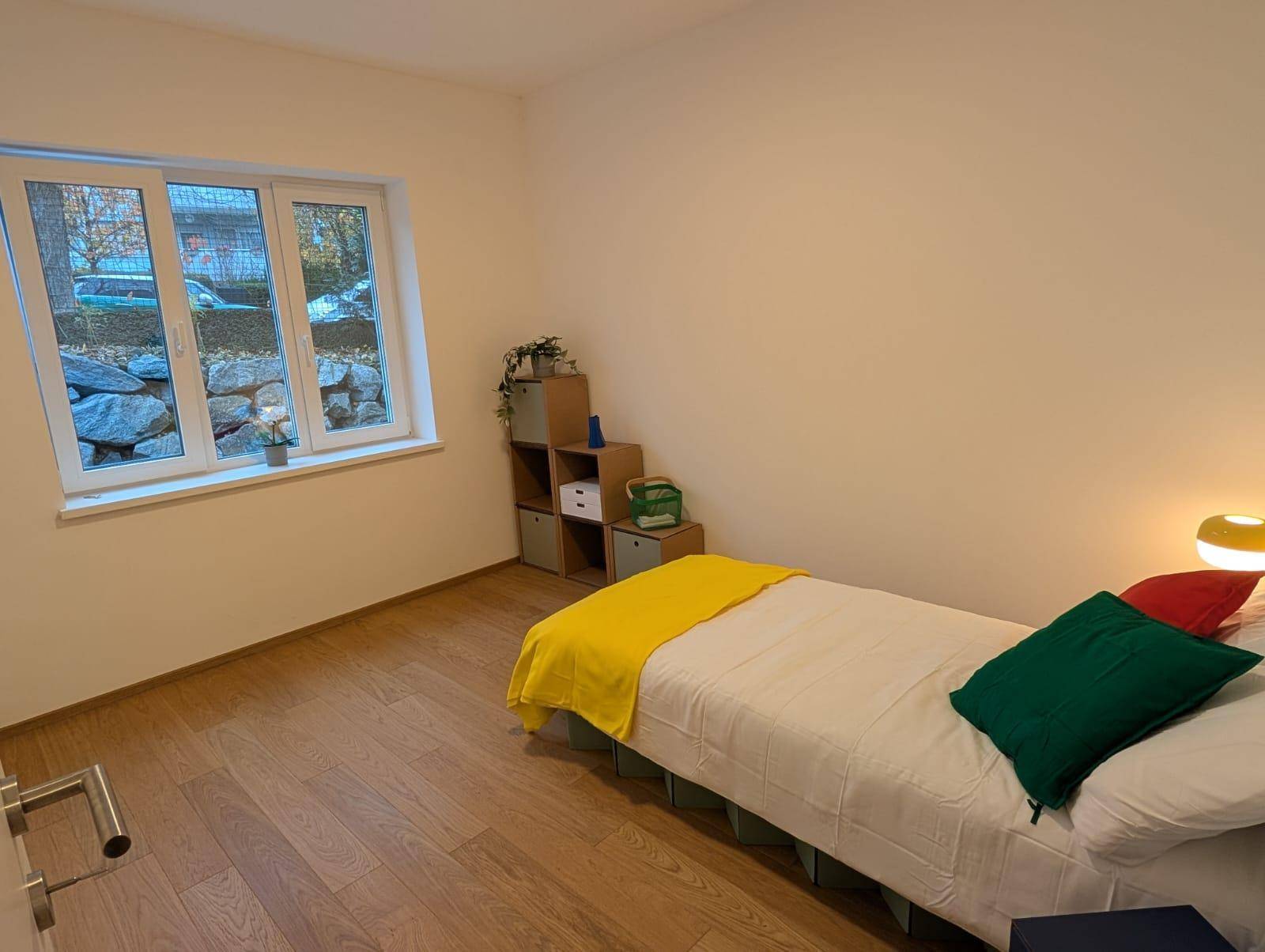 Schlafzimmer mit Holzboden, Fenster und Blick auf die Steinmauer im Außenbereich.