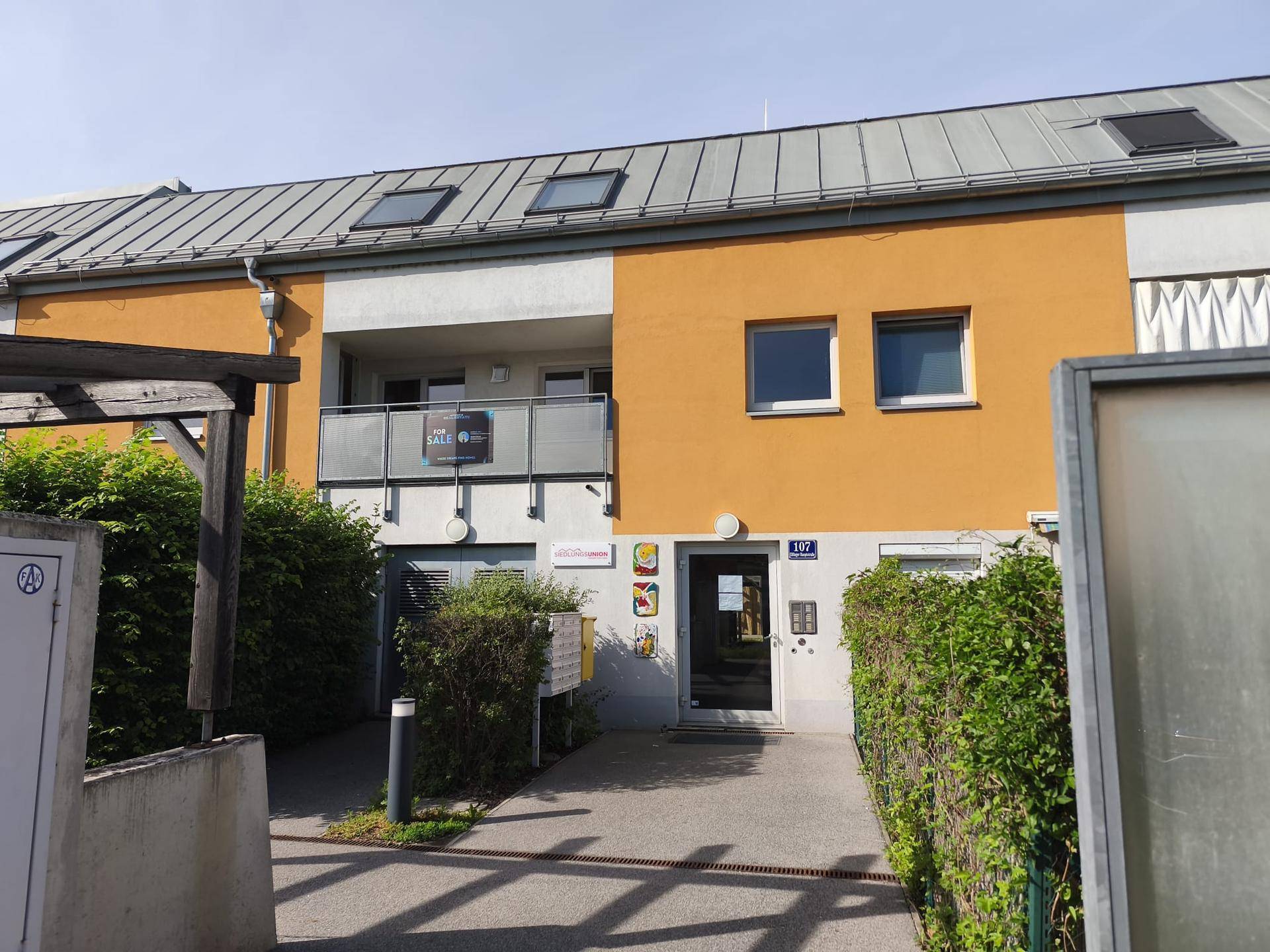 Eingangsbereich eines modernen Wohngebäudes mit orangefarbener Fassade und kleinem Balkon.