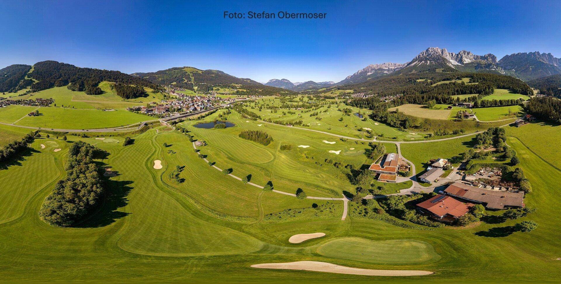 Luftaufnahme eines weitläufigen Golfplatzes mit angrenzendem Dorf und majestätischen Bergen im Hintergrund.