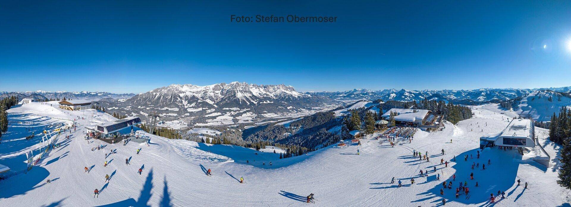 Weitläufige Winterlandschaft mit verschneiten Bergen, Skipisten und Skifahrern unter strahlend blauem Himmel.