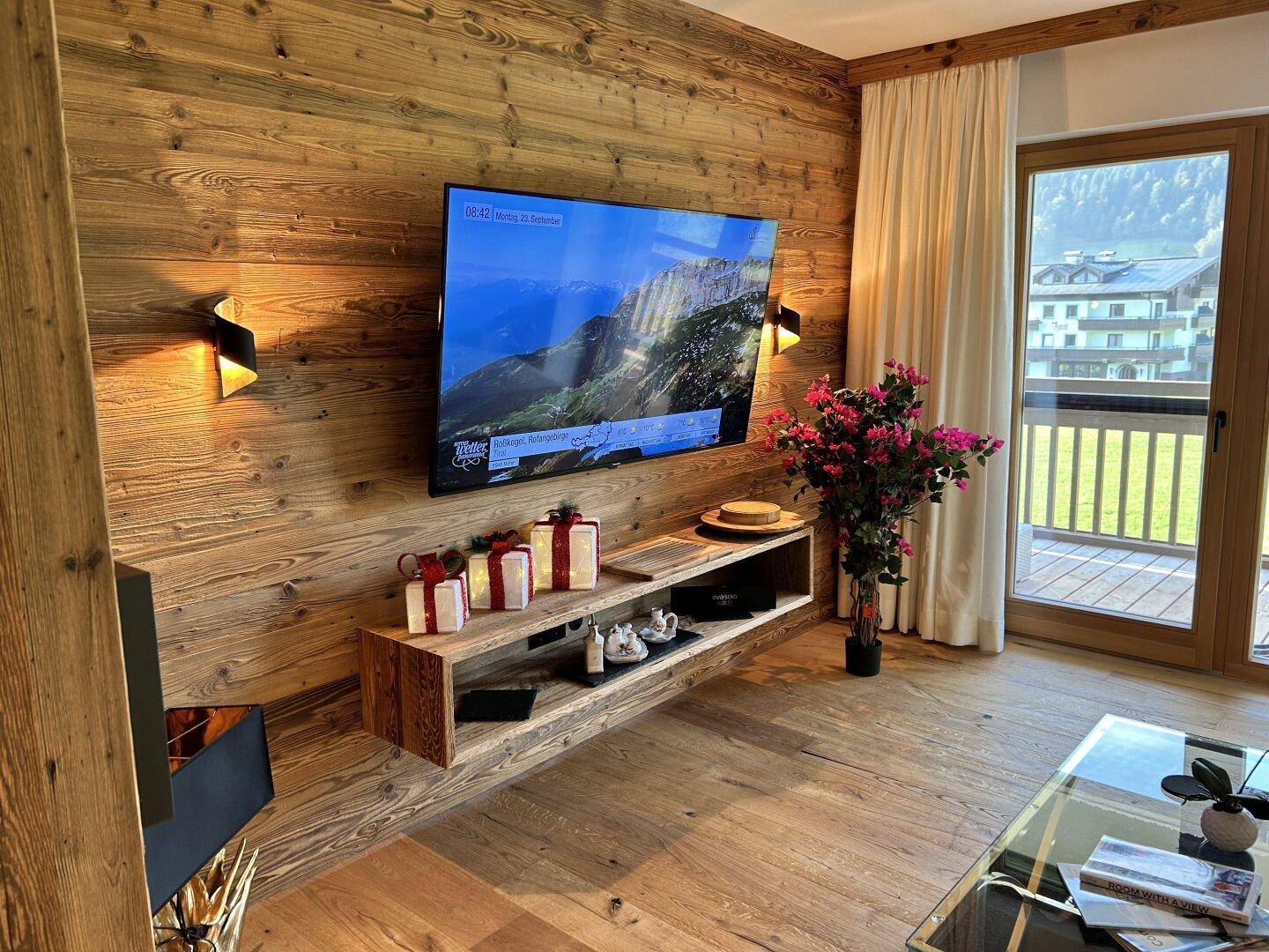 Wohnzimmer mit Holzwand, großem Fernseher und Zugang zu einem sonnigen Balkon mit Bergblick.