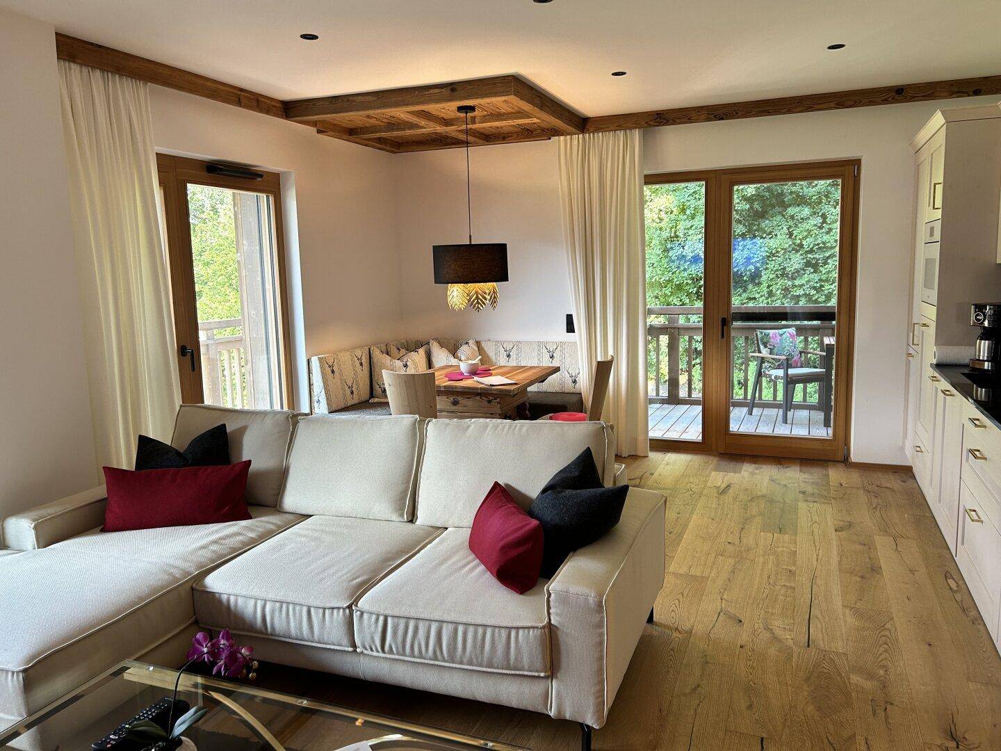 Offener Wohn-Essbereich mit beigem Sofa, Holzboden und Blick auf die grüne Umgebung durch große Fenster.