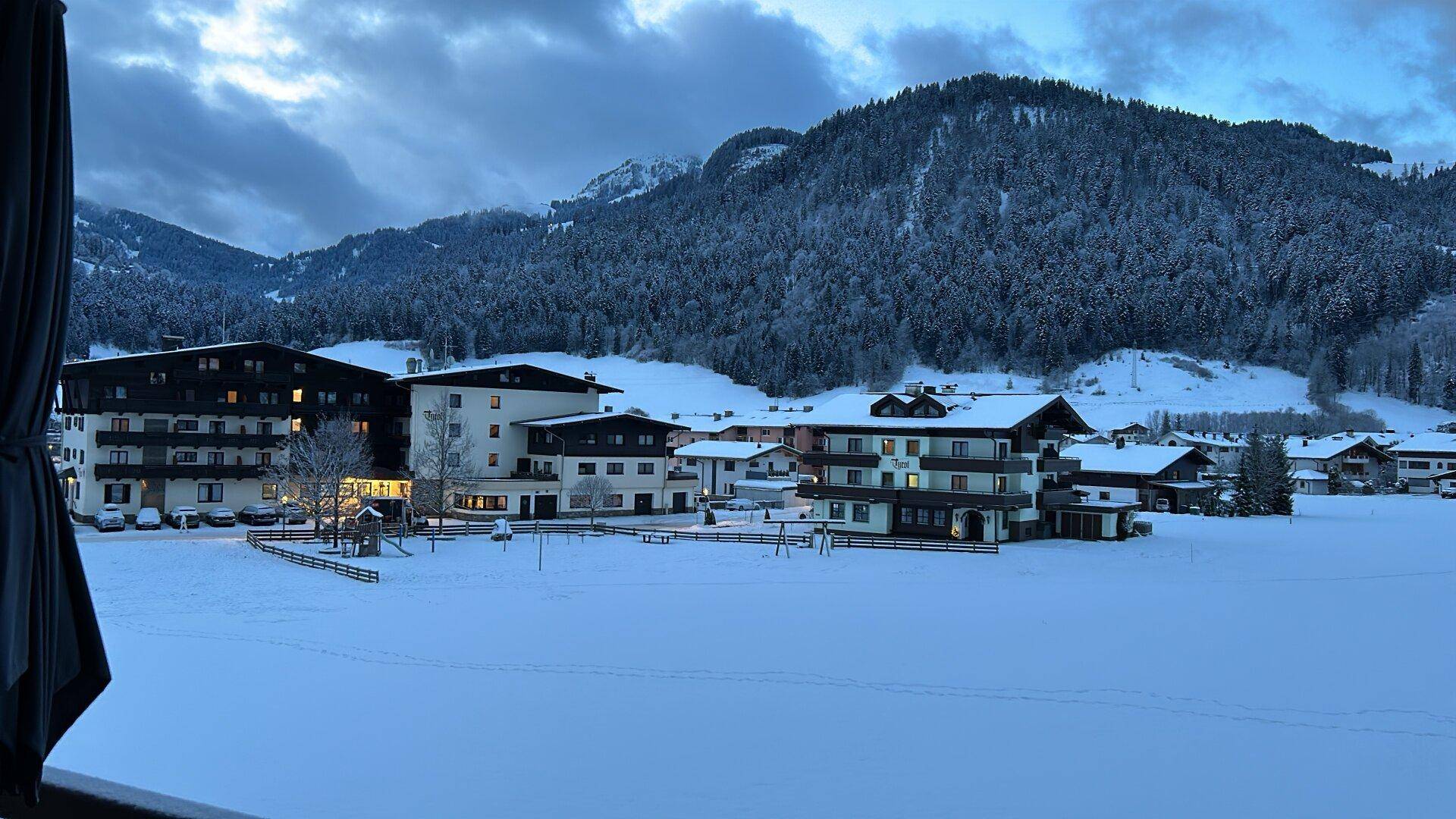 Winterliche Landschaft mit verschneiten Feldern, Bergen und umliegenden Gebäuden in der Dämmerung.