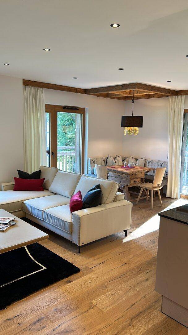 Heller Wohnbereich mit bequemer Couch, Holzboden und Essbereich im Hintergrund, mit Balkonzugang.