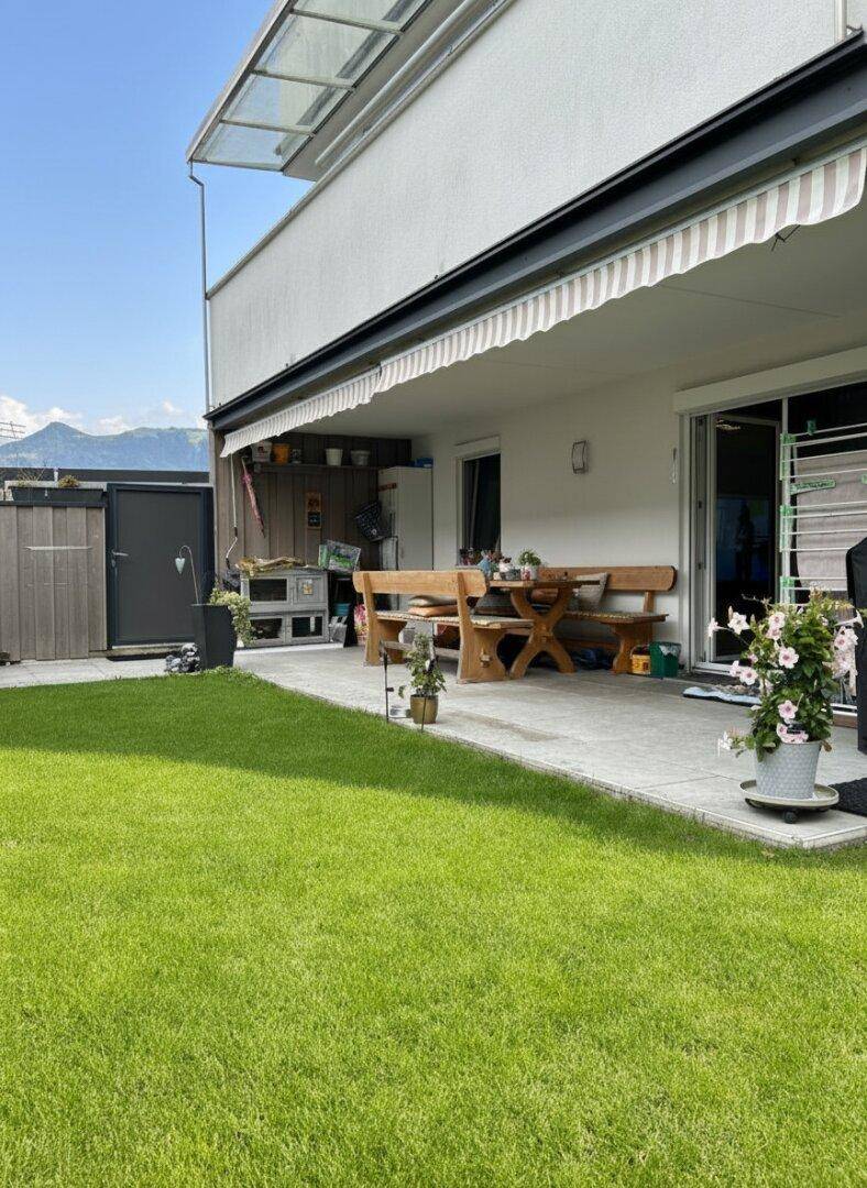 Sonnige Terrasse mit rustikalen Holzmöbeln, angrenzendem Rasen, Markise und Bergblick.