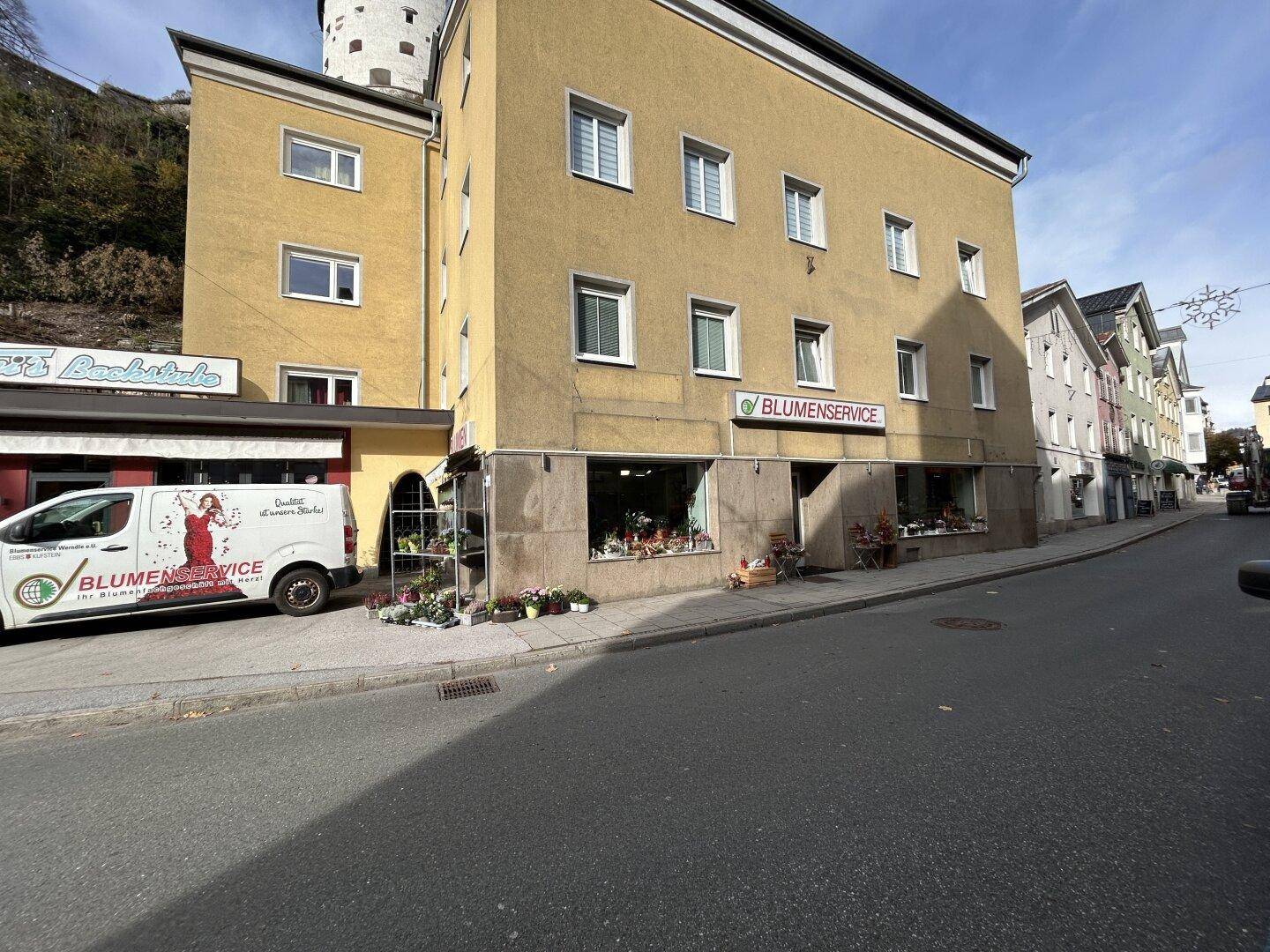 Gebäudeansicht von der Straße aus, mit dem Blumenladen im Erdgeschoss und dem historischen Schloss im Hintergrund.