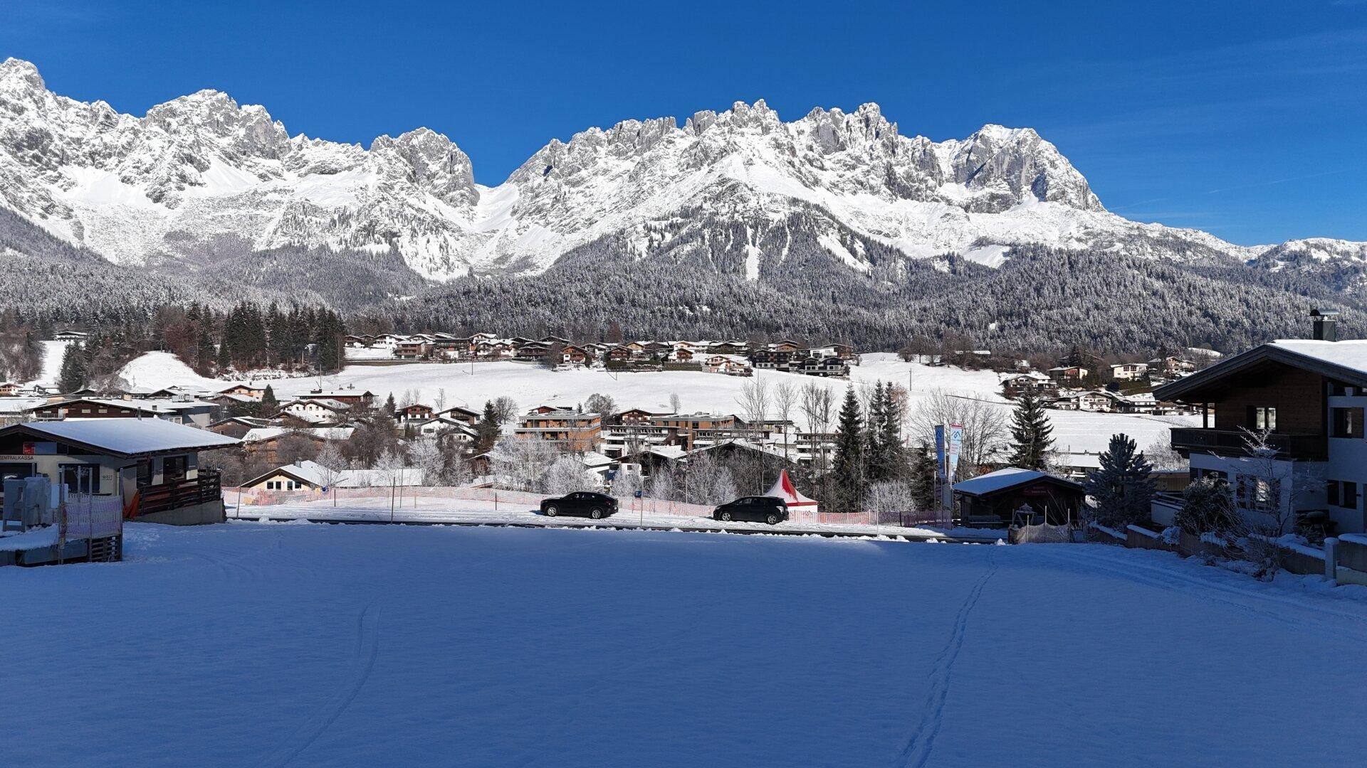 Panoramablick auf ein schneebedecktes Dorf mit imposanten, schneebedeckten Bergen im Hintergrund unter blauem Himmel.