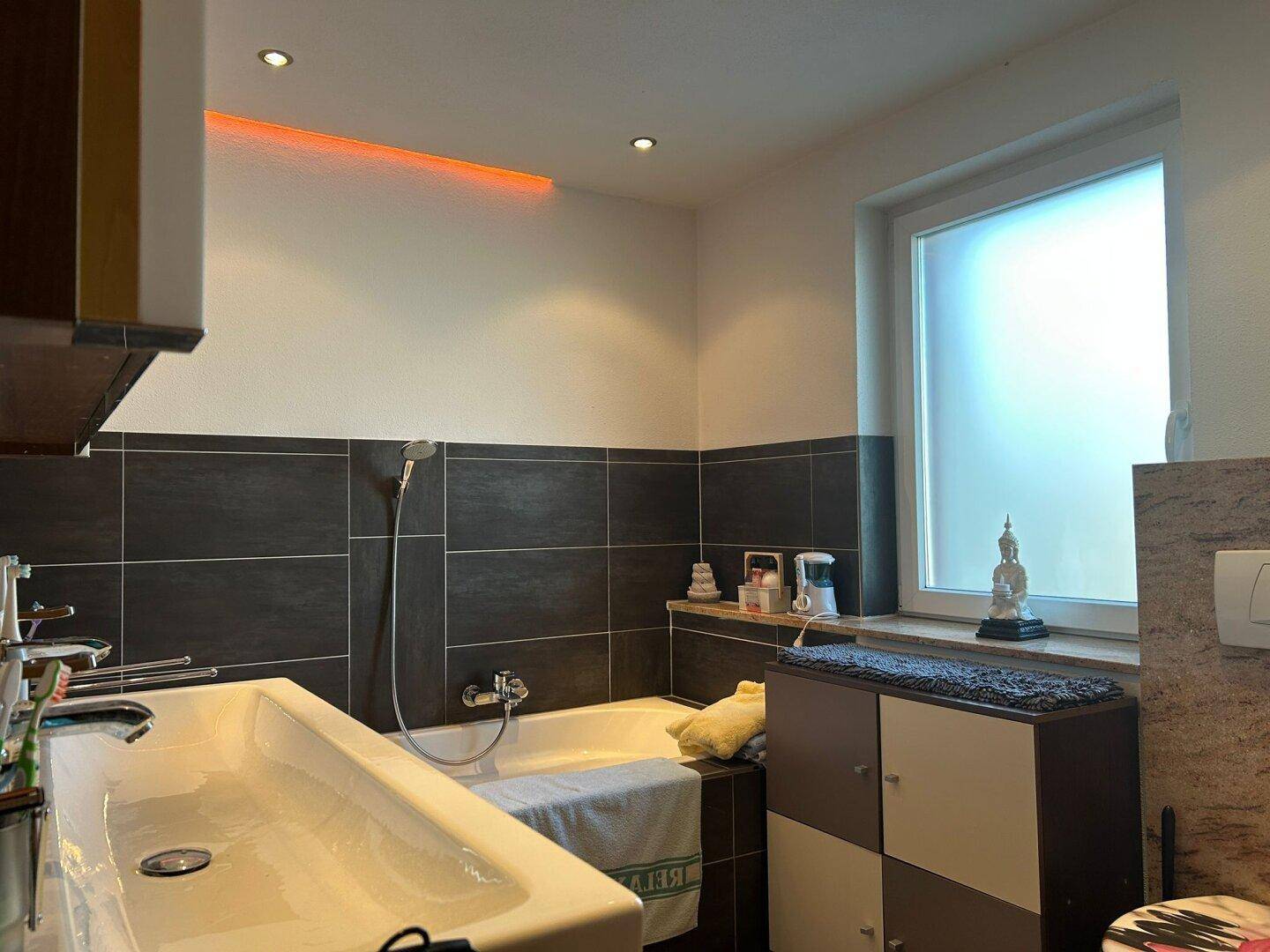 Modernes Badezimmer mit Badewanne, dunklen Fliesen und indirekter Beleuchtung, sowie einem Fenster.