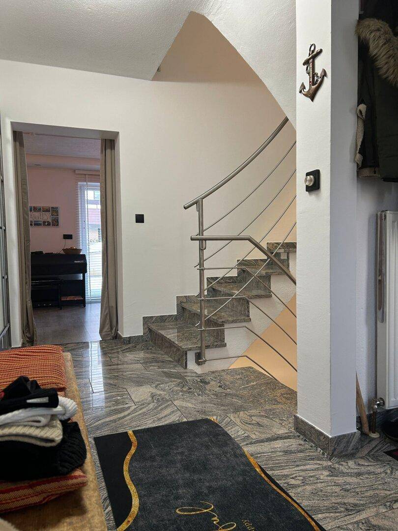 Heller Flur mit Steintreppe, Edelstahlgeländer und gemustertem Boden, der zum nächsten Stockwerk führt.