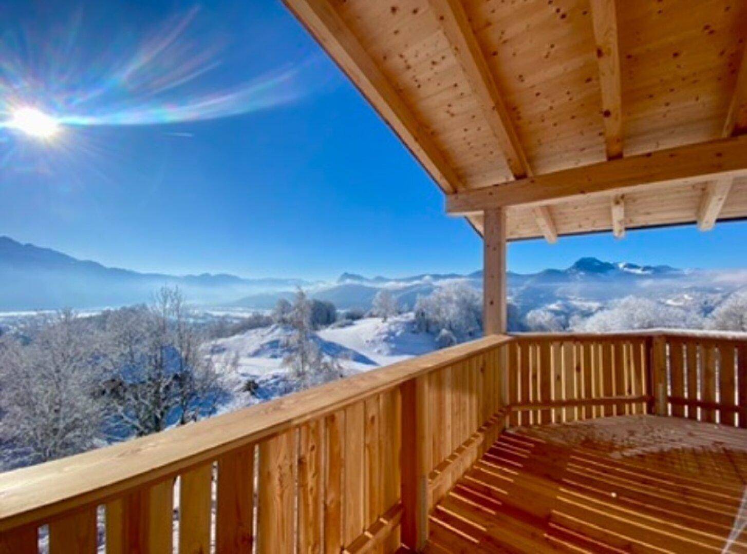 Sonniger Holzbalkon mit weitem Blick über eine verschneite Winterlandschaft und Berge.