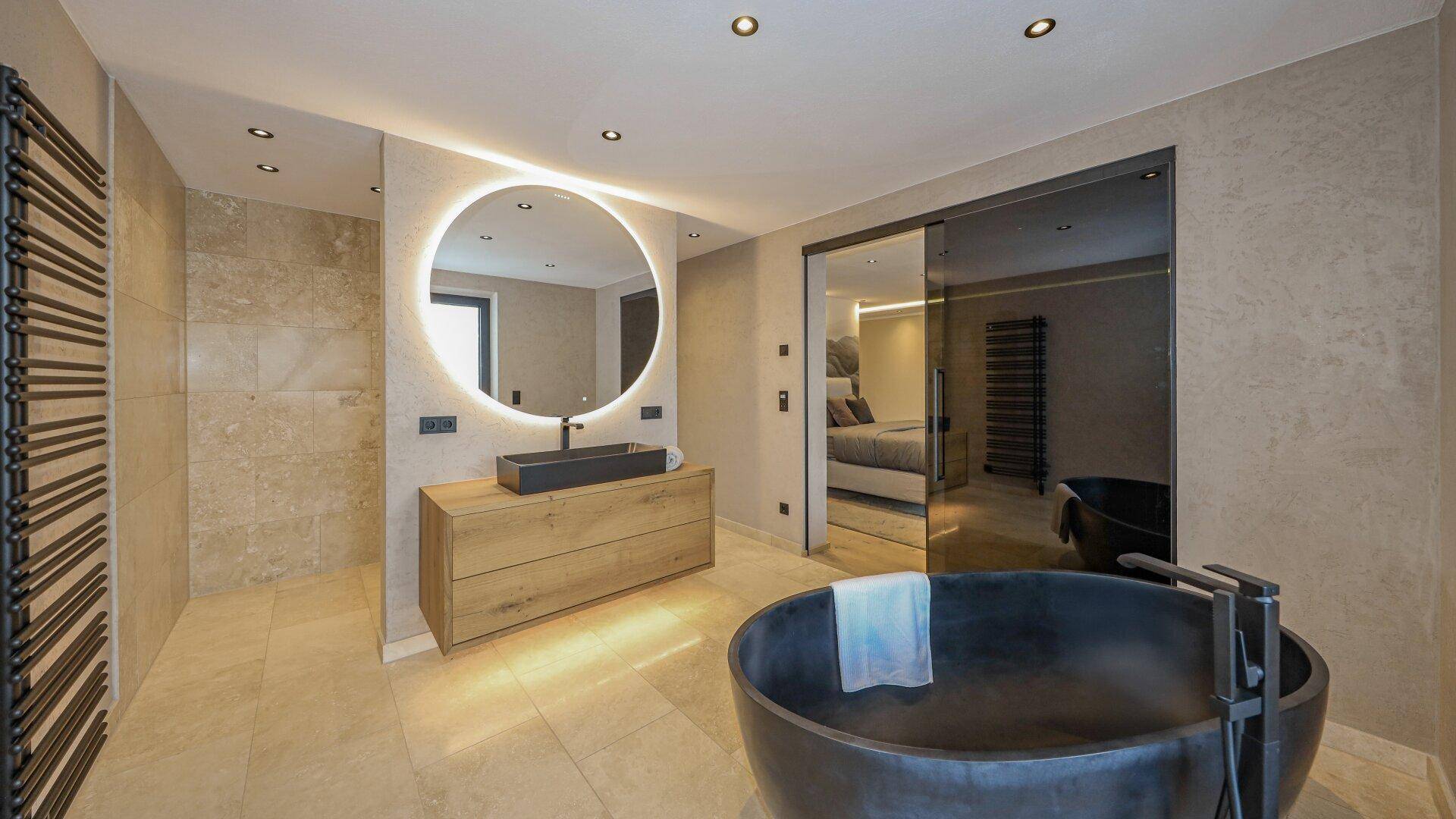 Luxuriöses Badezimmer mit freistehender Badewanne, beleuchtetem Spiegel und Natursteinfliesen.