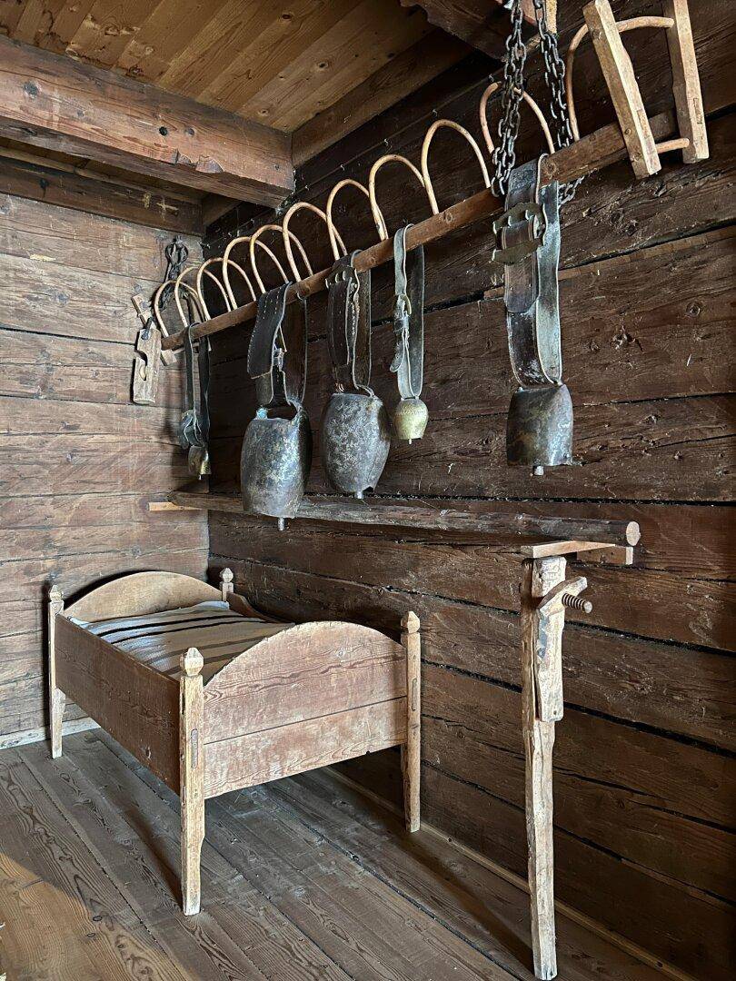 Rustikaler Raum mit Holzwänden, einem kleinen Holzbett und traditionellen Kuhglocken als Dekoration.