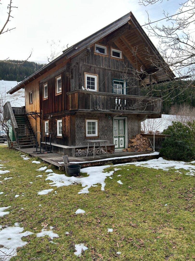 Traditionelles Holzhaus mit Balkon und Außentreppe, umgeben von einer teilweise verschneiten Wiese.