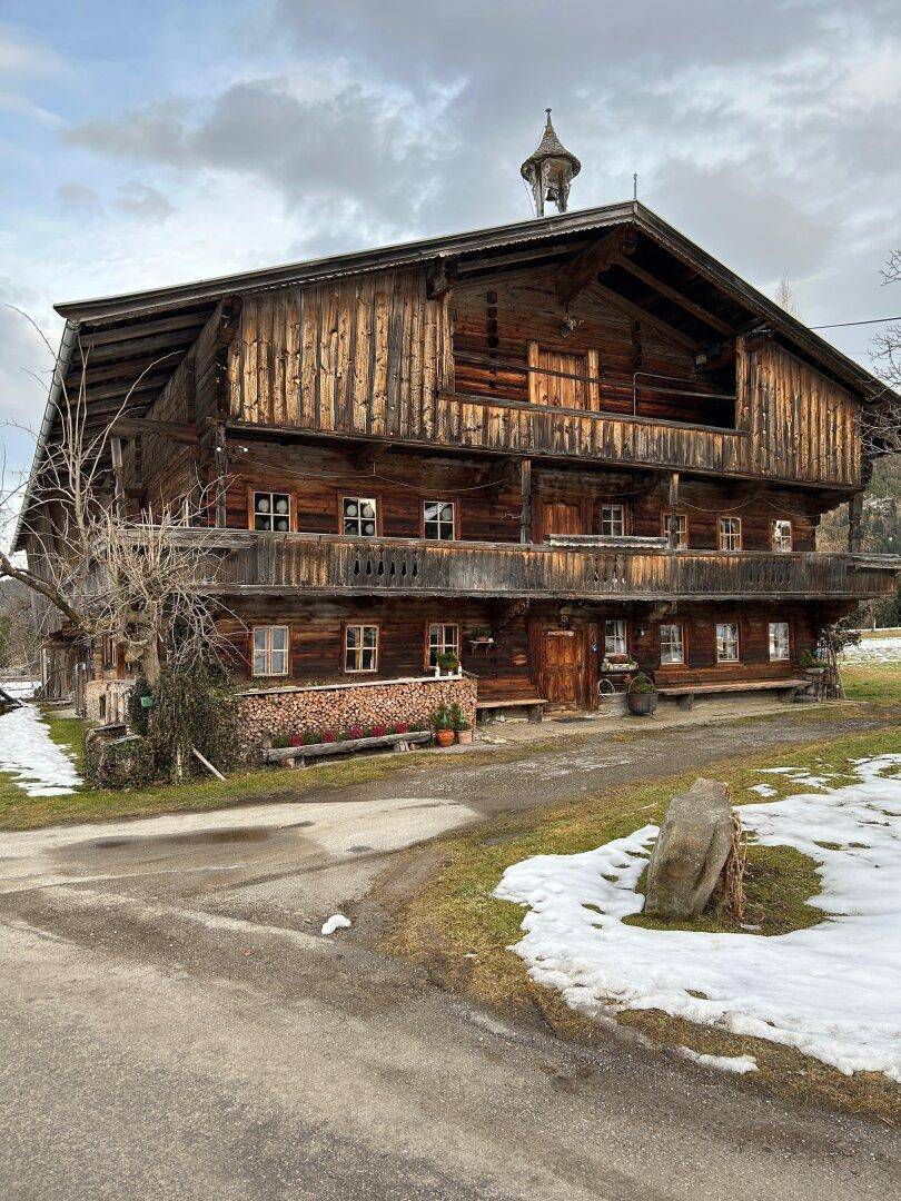 Traditionelles Holzhaus mit mehreren Balkonen und einem Glockenturm, umgeben von leicht verschneiter Landschaft.