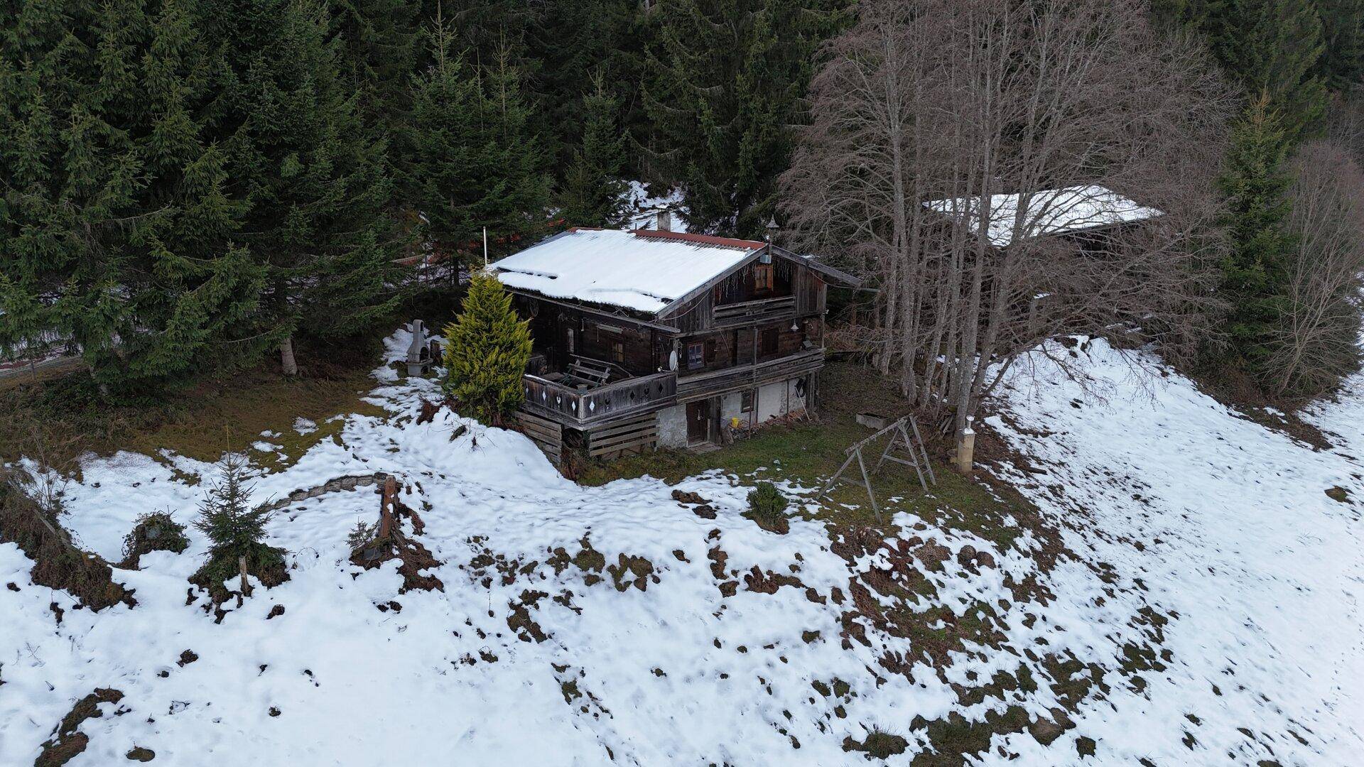 Traditionelles Holzhaus auf einem Hang, umgeben von Wald und verschneiten Feldern.