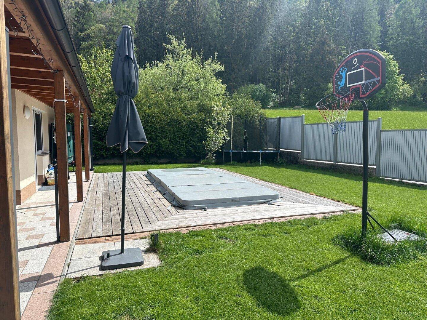 Sonniger Garten mit Poolabdeckung, Trampolin und Basketballkorb, umgeben von Grün.
